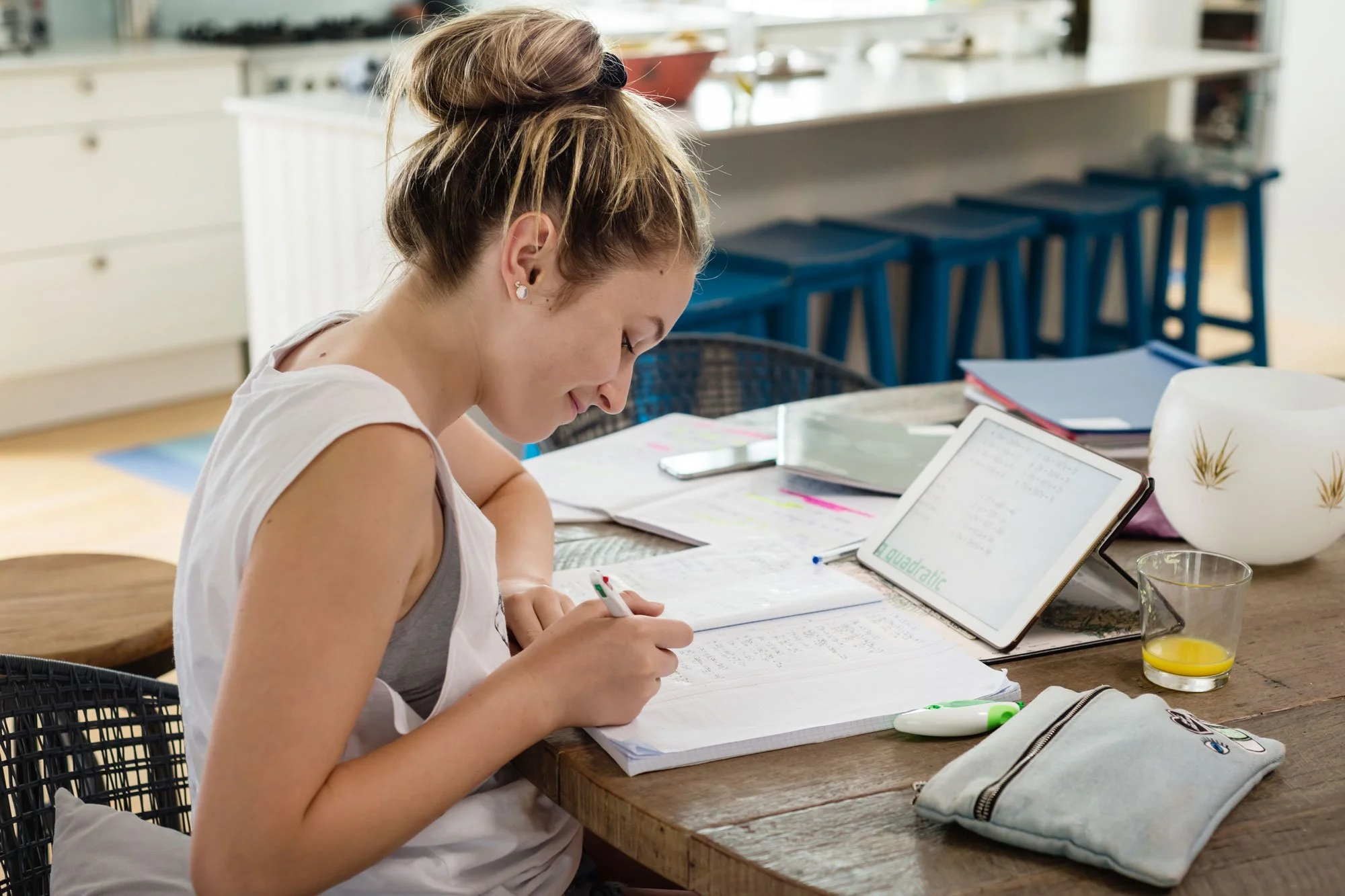 A young woman is sitting at a wooden table, writing in a notebook with a marker, surrounded by study materials in a bright kitchen. There is an iPad with educational content, glasses of drinks, a pencil case, and other stationery items on the table.