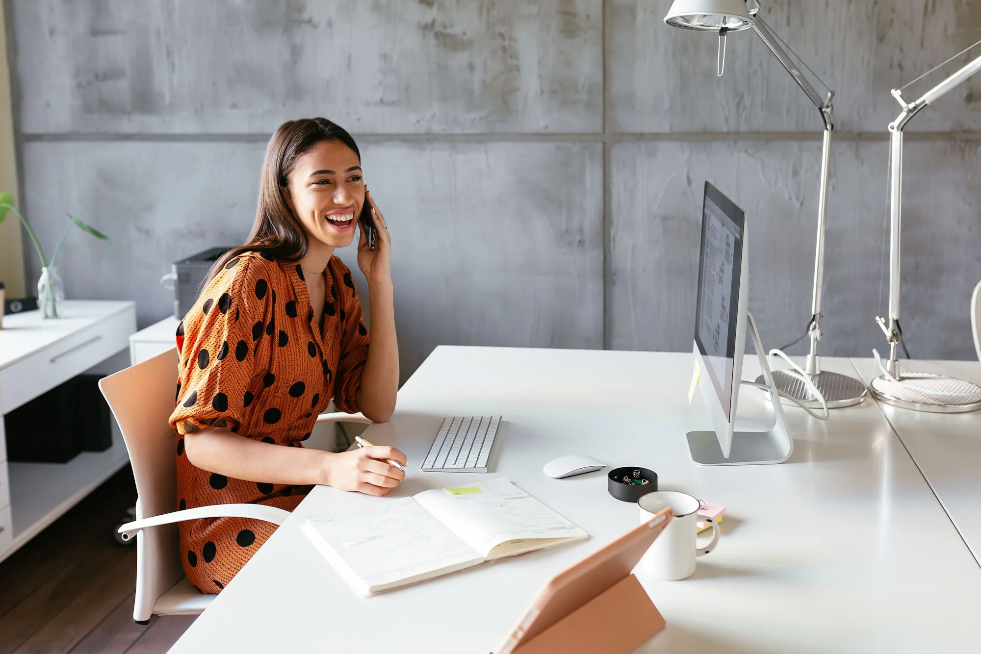 A woman sitting at a white desk, talking on her cell phone, smiling, with a computer, keyboard, mouse, open notebook, cups, and desk lamp in a modern office.
