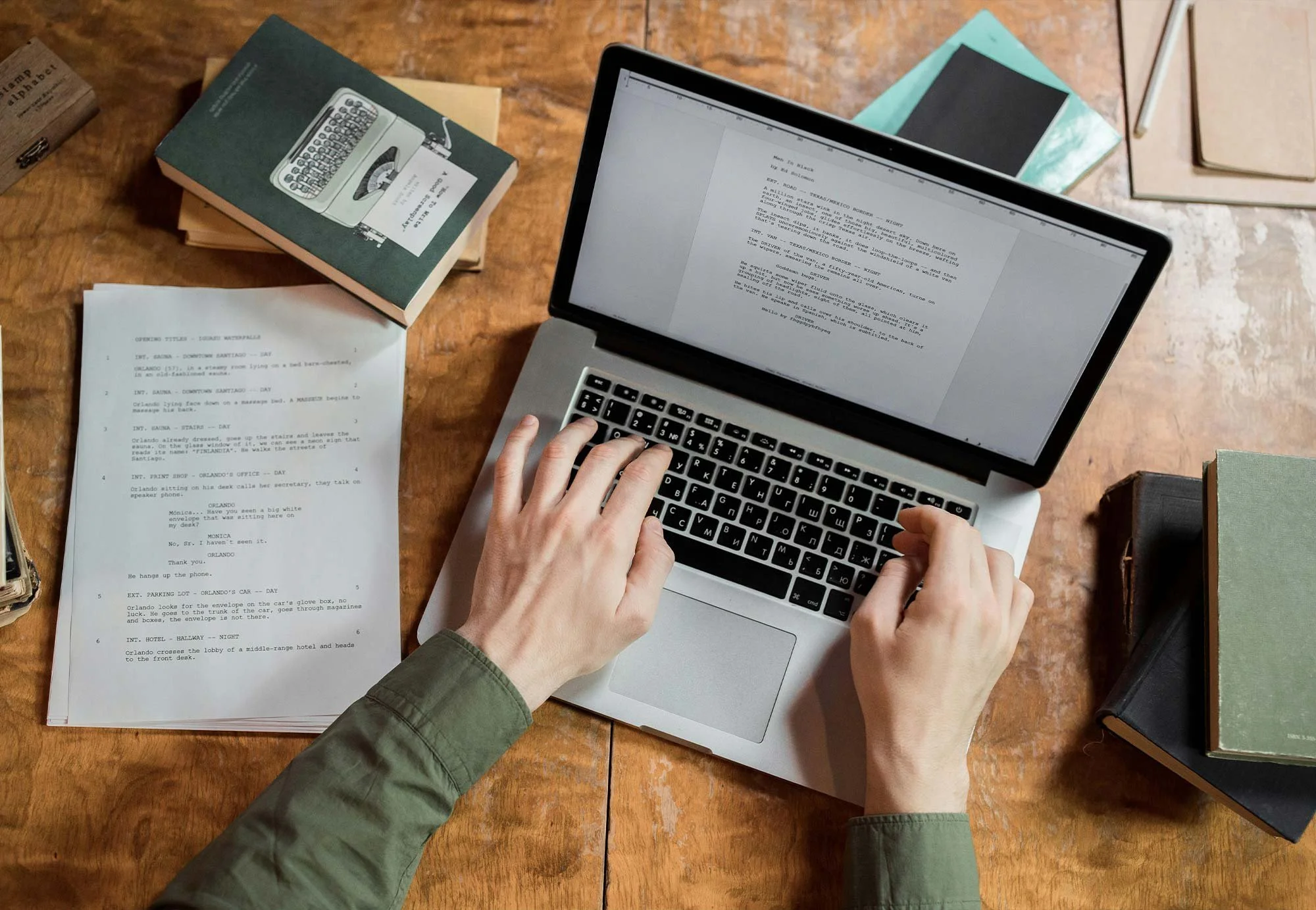 Person typing on a silver laptop with a screen displaying a script, surrounded by printed pages, books, and notebooks on a wooden table.