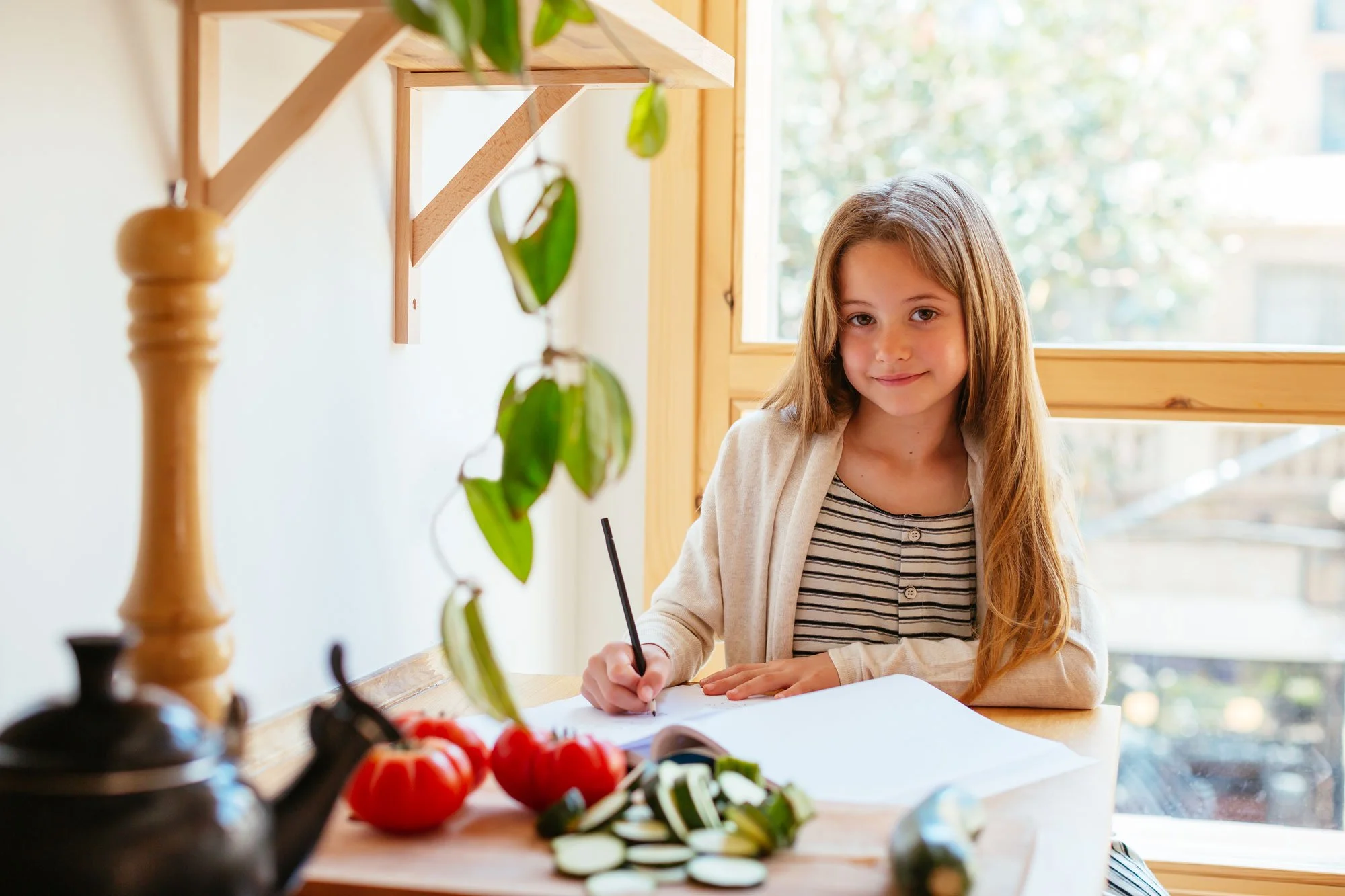 A young girl with long light brown hair sitting at a wooden table by a window, holding a black pen, with tomatoes and sliced vegetables on the table, and a green leafy plant in the foreground.