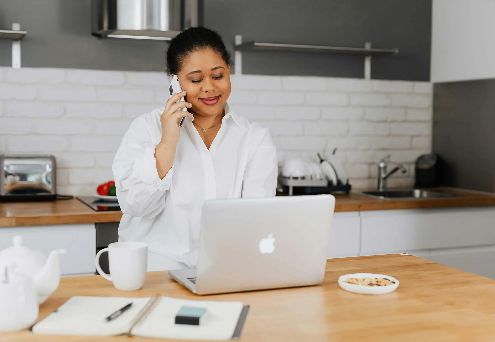Woman talking on the phone while working on a laptop in a modern kitchen.