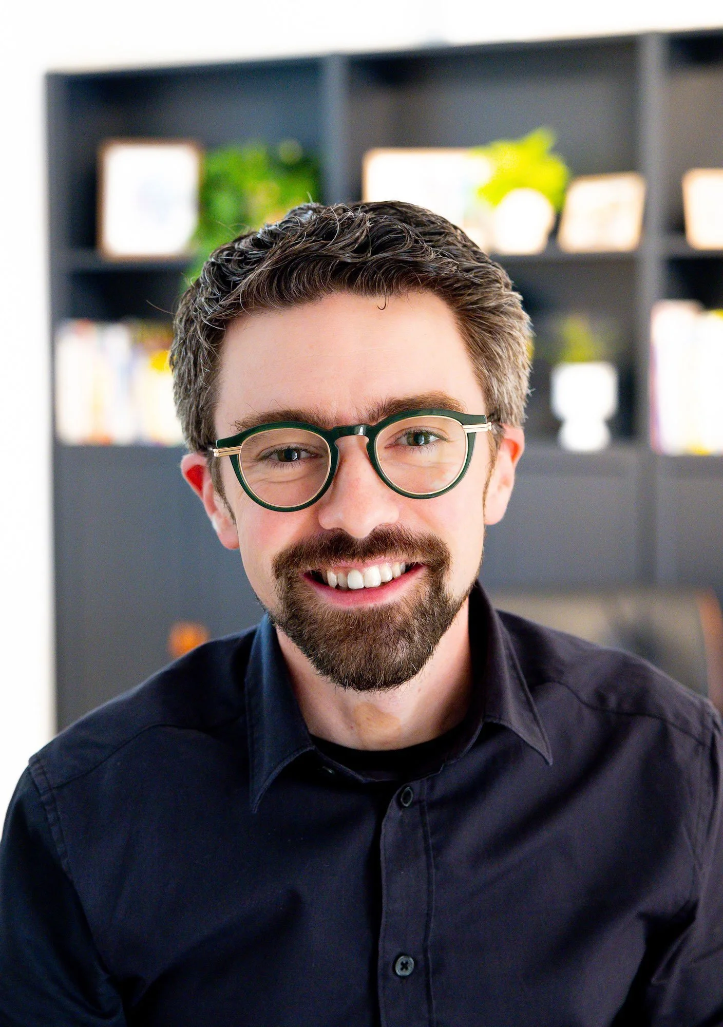 A smiling man with glasses and a beard, wearing a black shirt, in front of a bookshelf with plants and books.