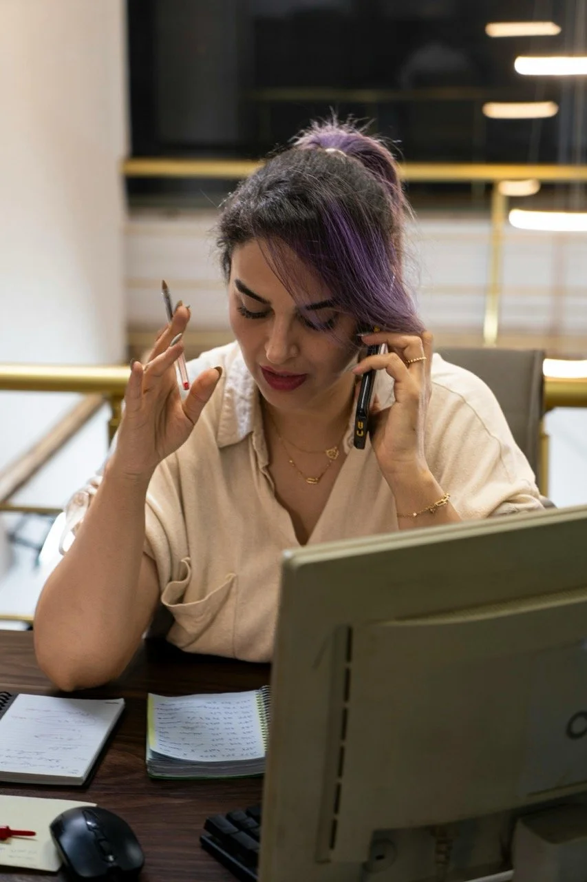 A woman with dark hair and purple streaks, wearing a beige shirt, talking on the phone while holding a pen, sitting at a desk with notebooks and a computer monitor.