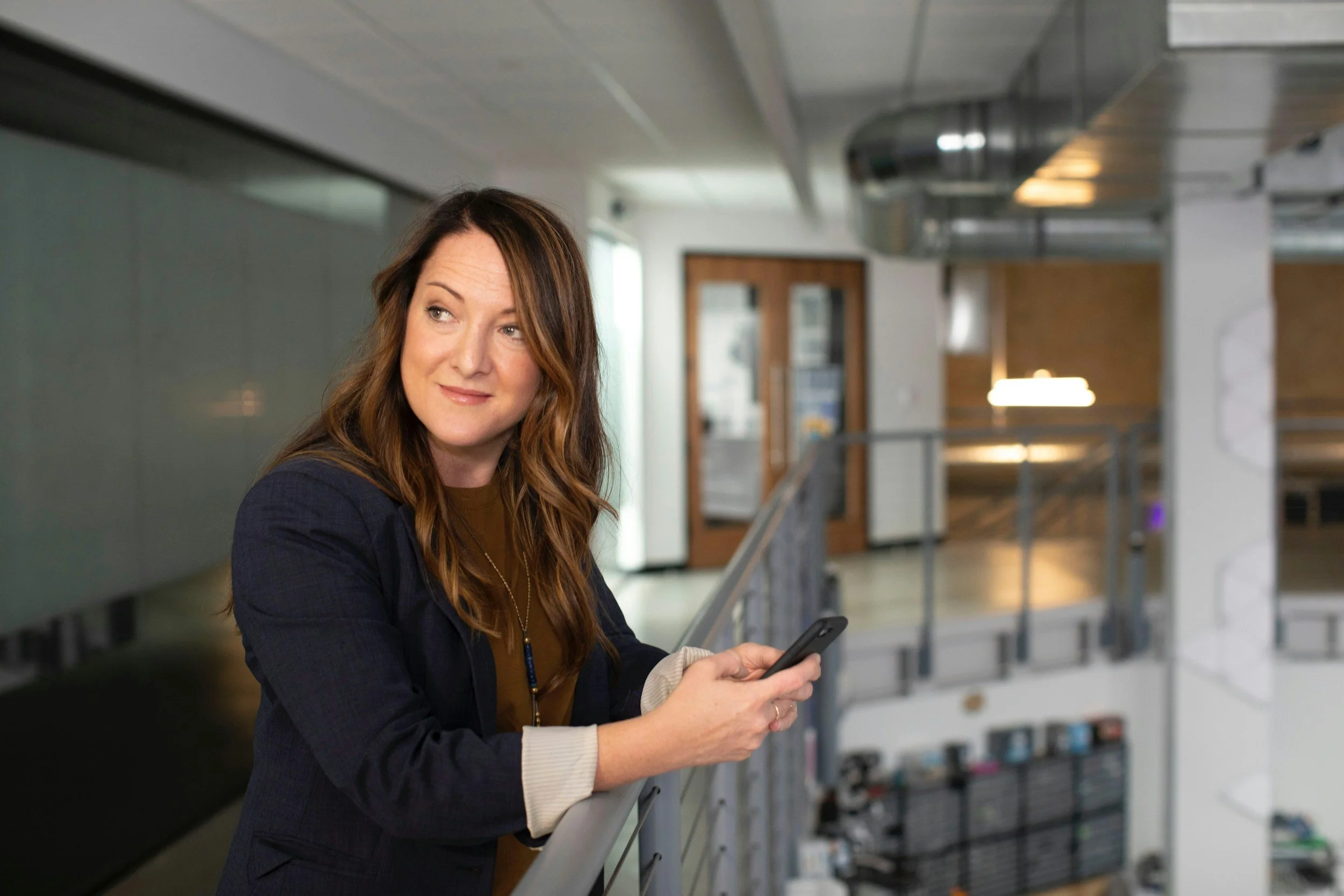 A woman with long red hair and a dark blazer leaning on a railing in a modern indoor space, holding a smartphone, with a background of a staircase and bright natural light.