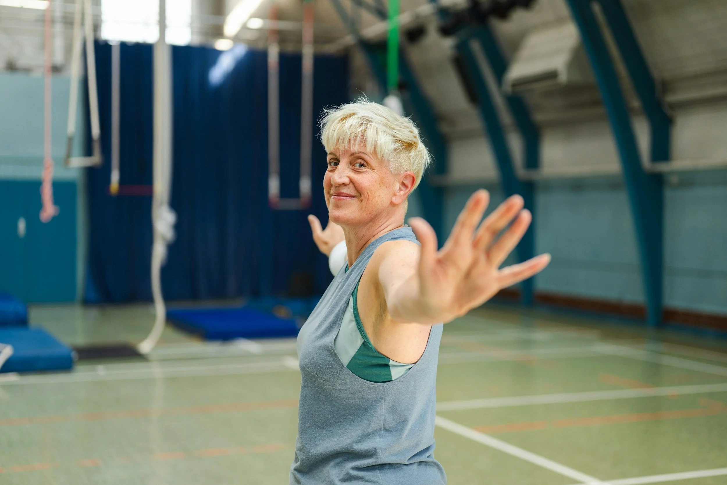 An older woman with short blonde hair smiling and extending her arms in a gymnasium.