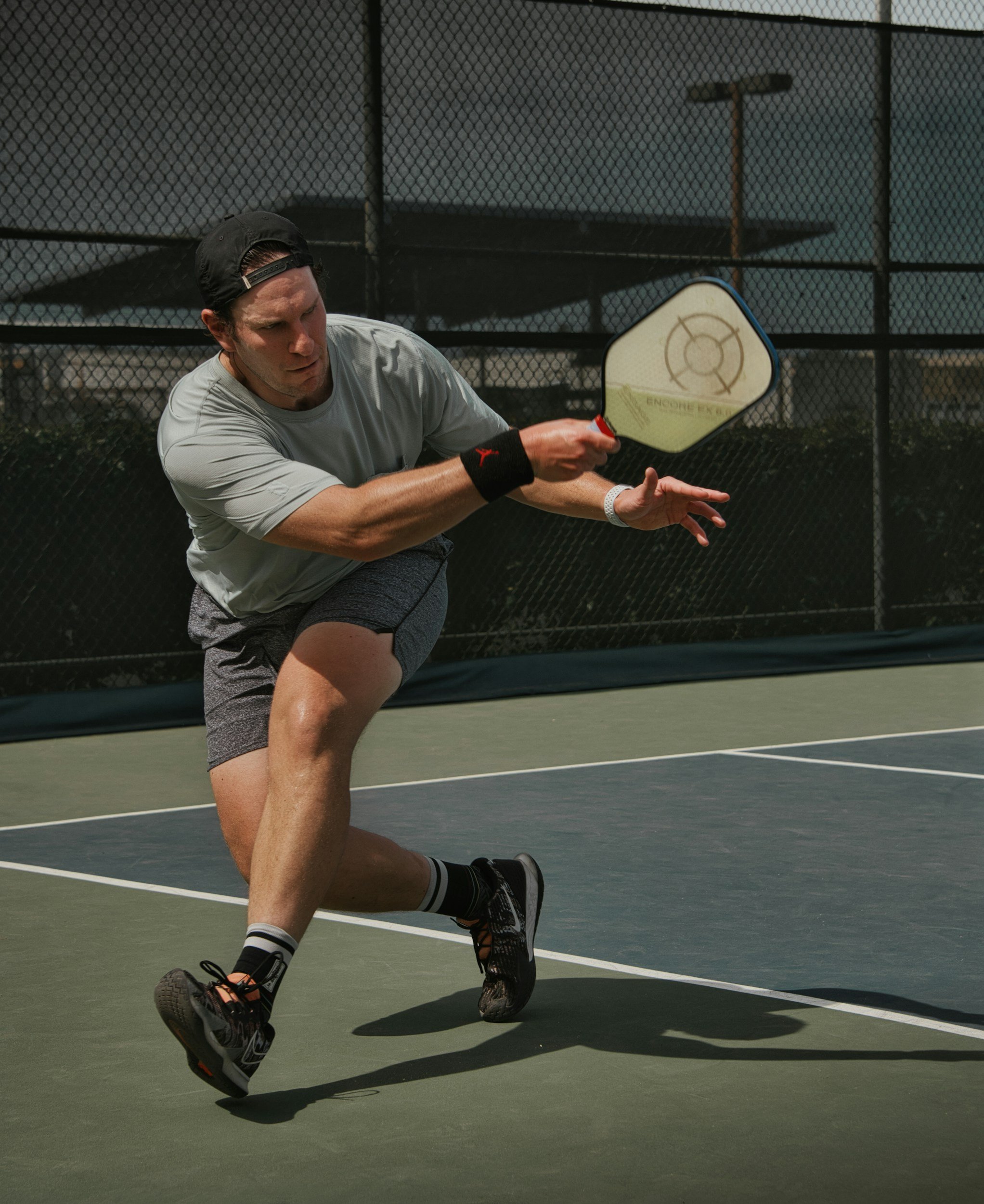 A man playing pickleball on an outdoor court, wearing a gray T-shirt, black cap, black wristband, shorts, and sneakers, preparing to hit a ball with a paddle.