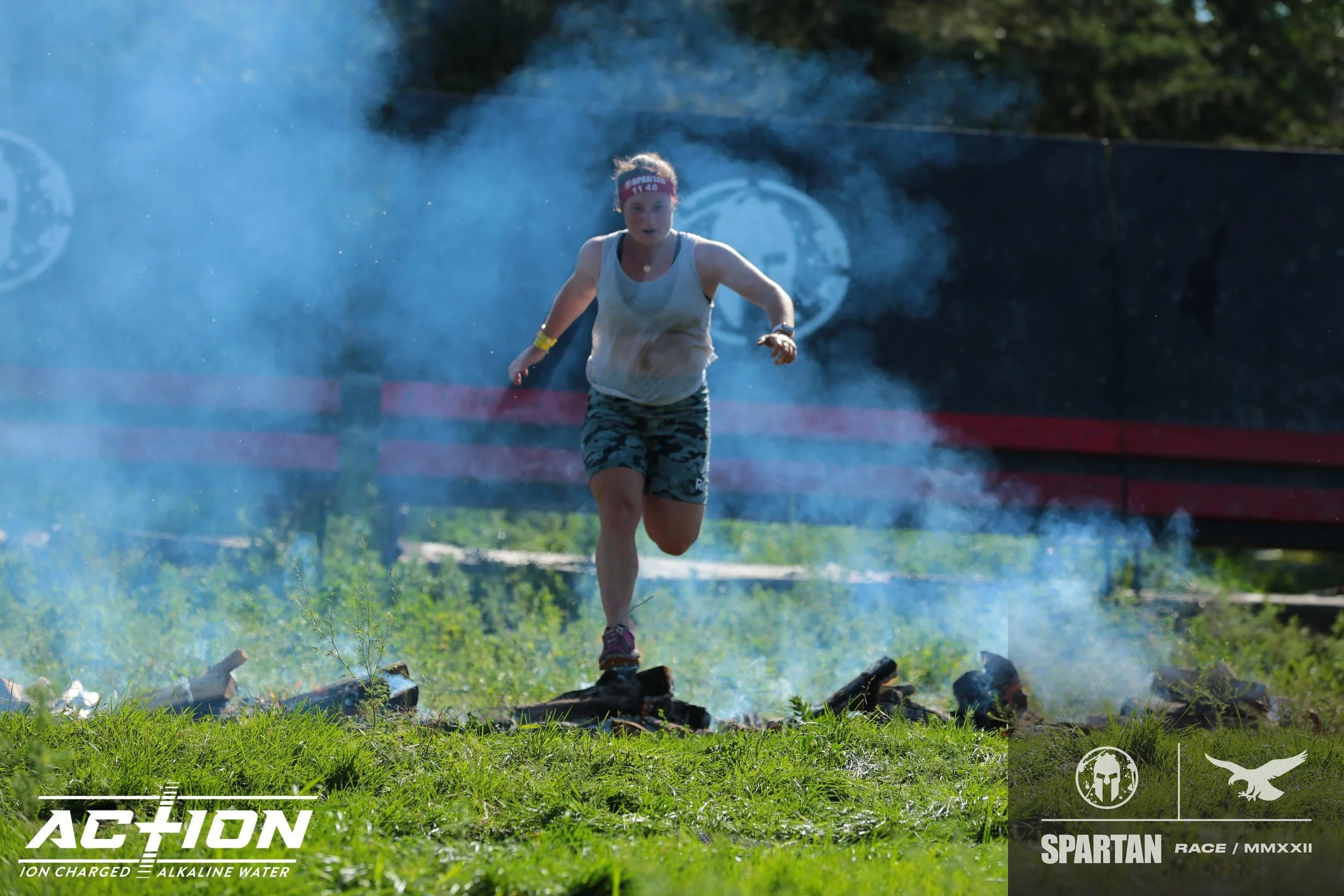 Female participant running on an obstacle course during a Spartan race, surrounded by smoke, with a black fence and green trees in the background.