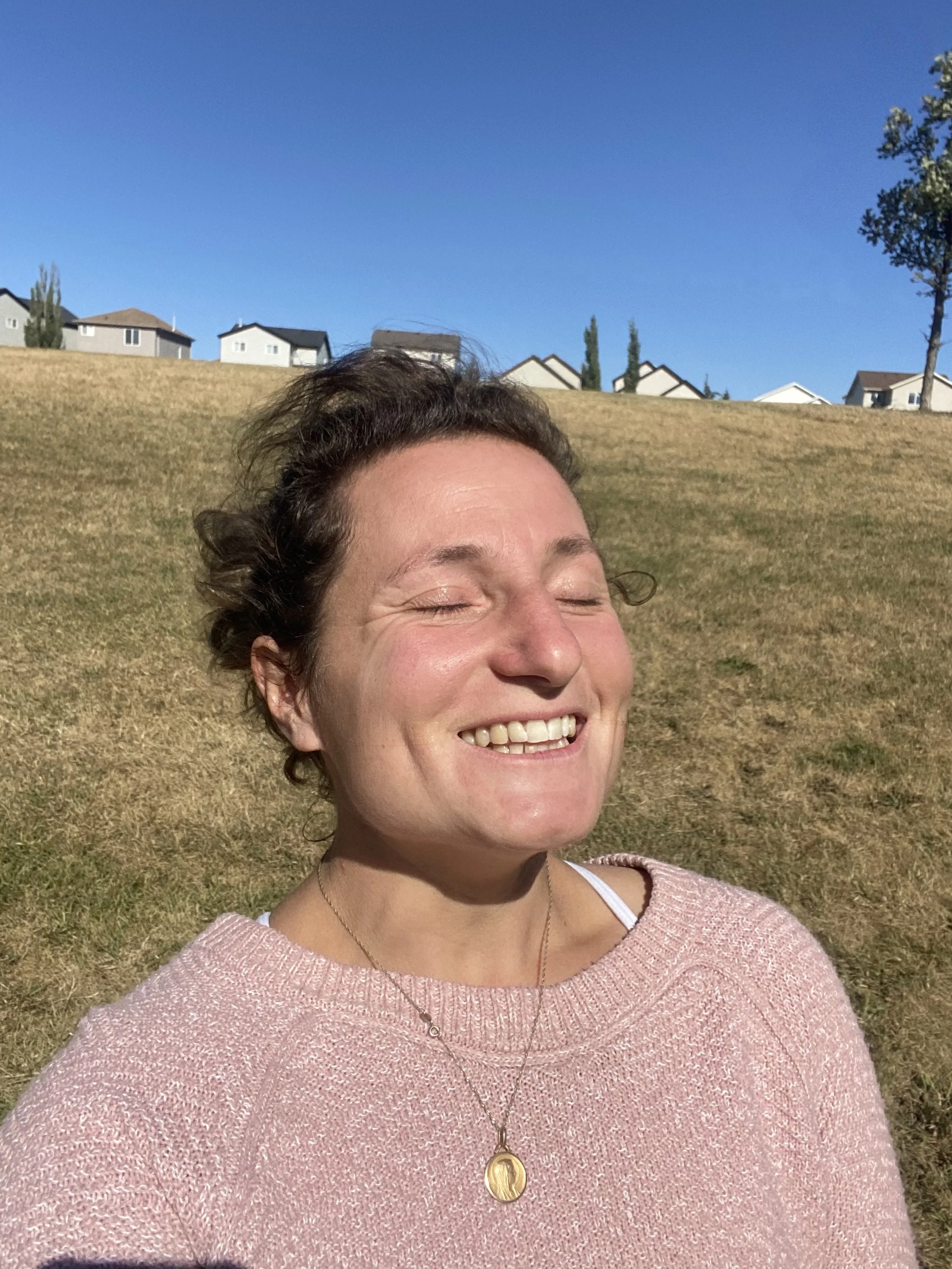 A woman smiling with eyes closed outdoors on a sunny day, with a grassy hillside and suburban houses in the background.