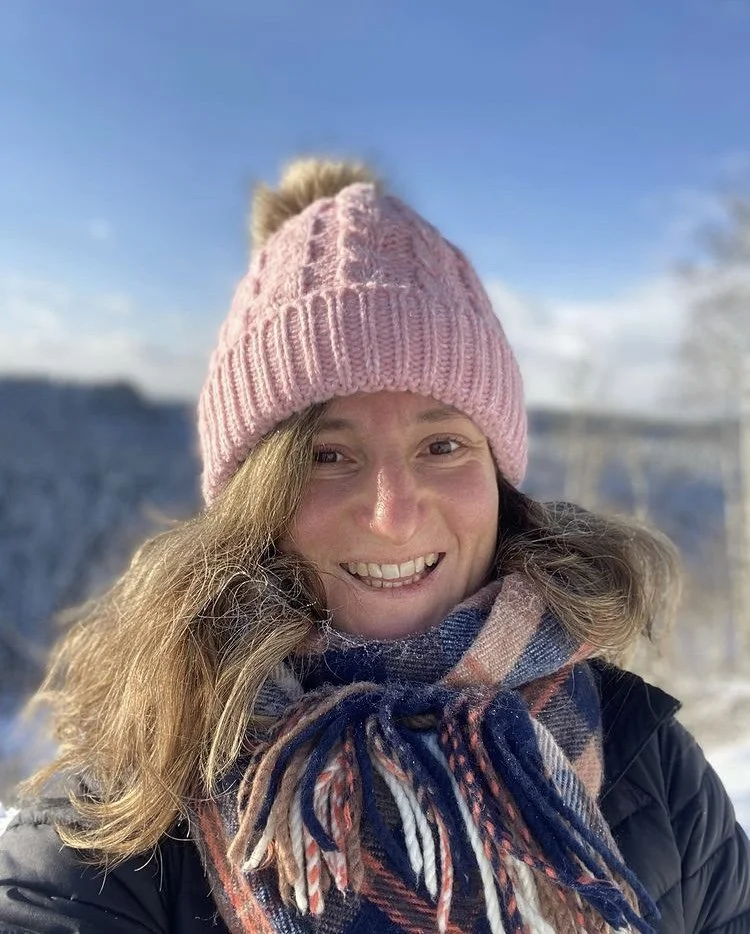 A woman smiling in winter outdoors, wearing a pink knit beanie with a pom-pom and a colorful striped scarf.