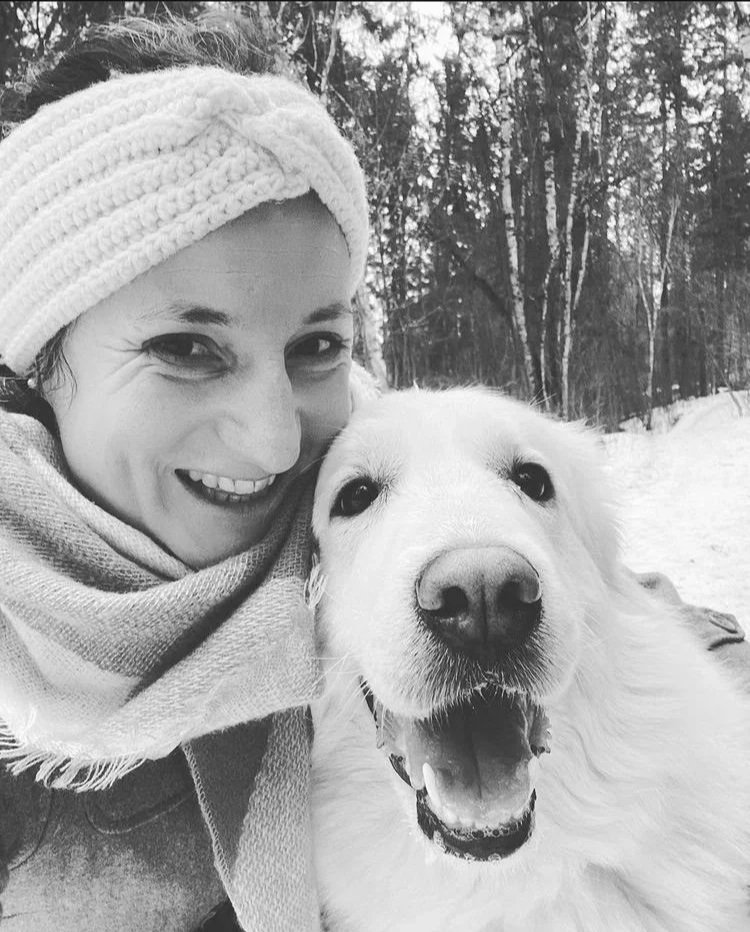 A woman with a headband and scarf smiling next to a happy golden retriever dog in a snowy outdoor setting with trees in the background.