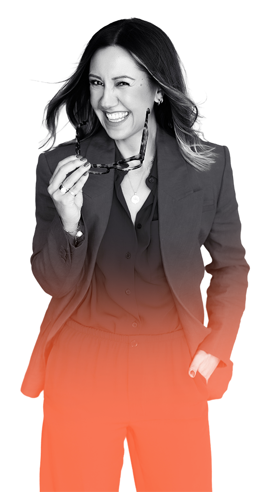 Kate Dillon smiling and winking, holding sunglasses, dressed in a blazer and pants.