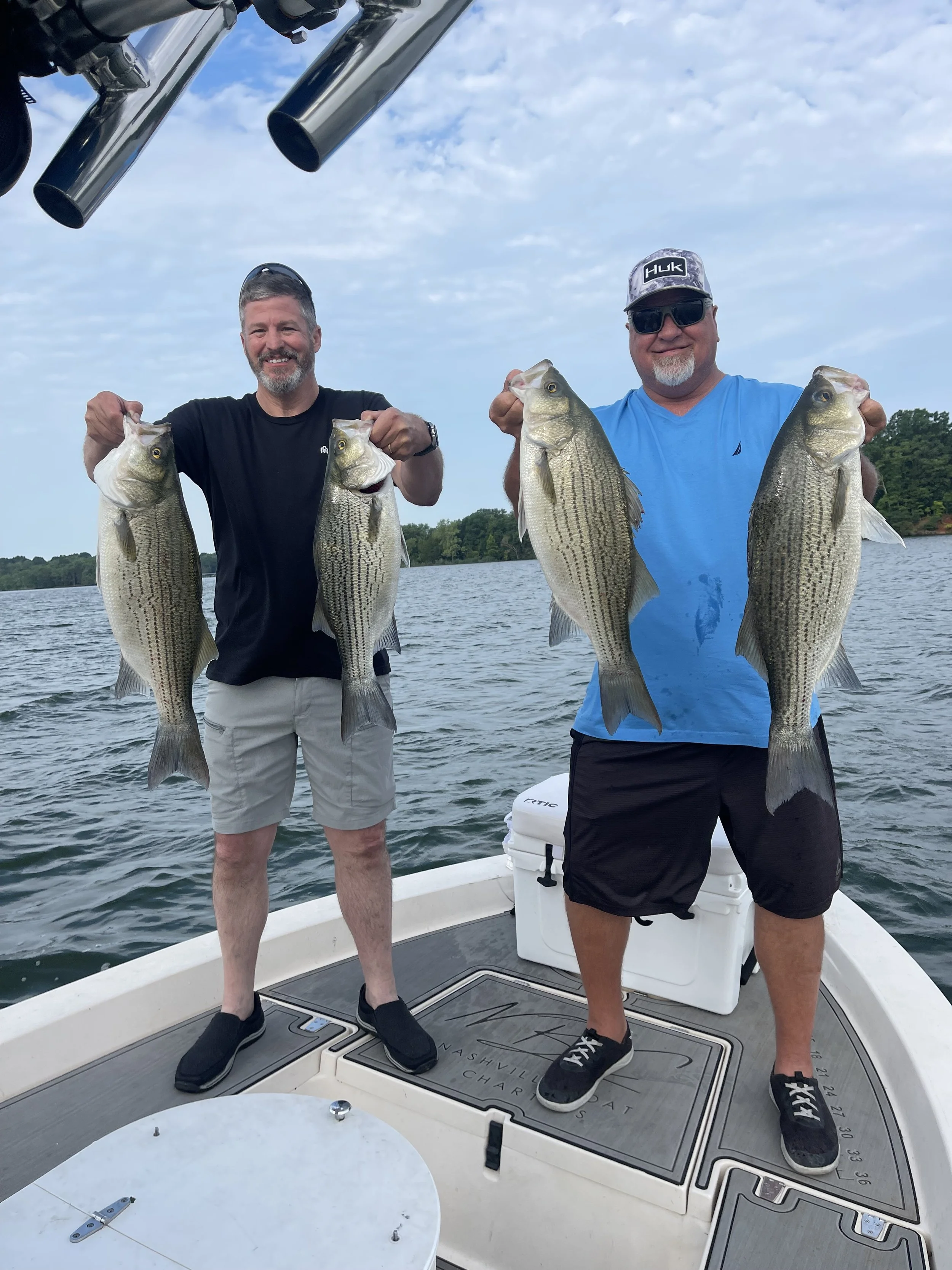 Two men on a boat holding four striped bass fish, smiling, with water and trees in the background.