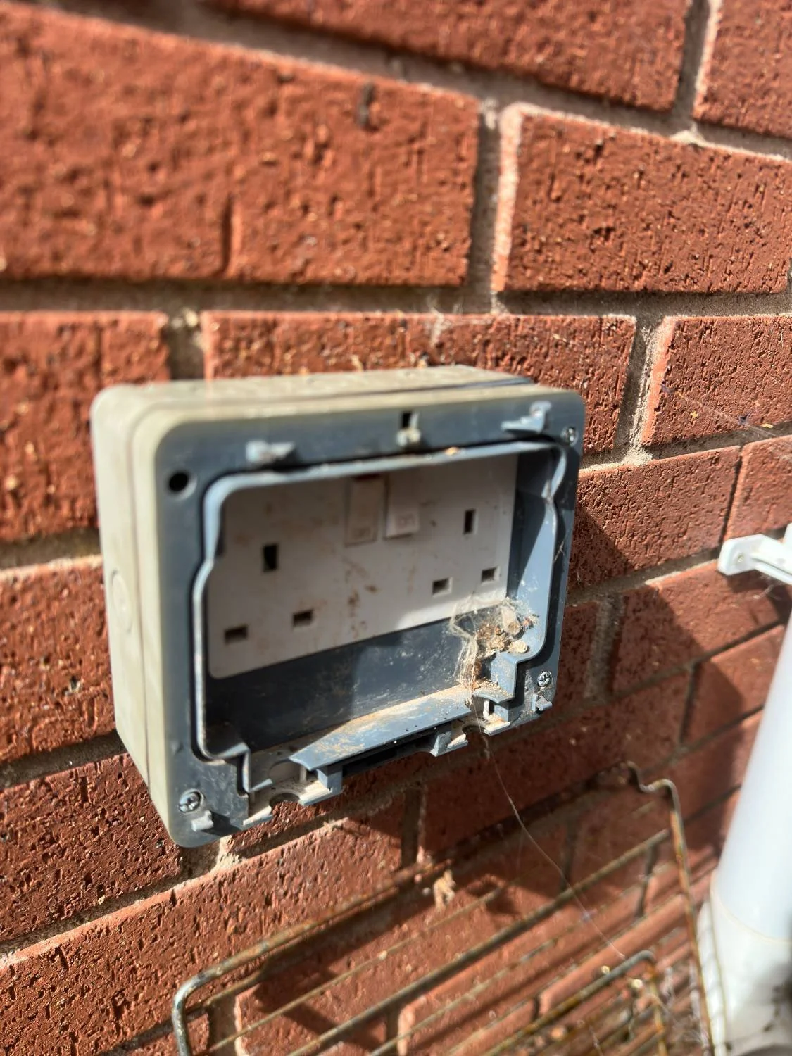 An old, damaged, empty electrical outlet mounted on a brick wall with some dust and spider webs.