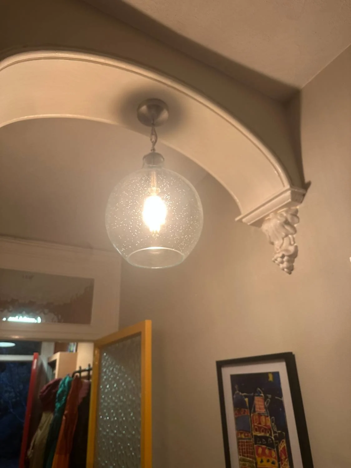 Ceiling light fixture with a clear glass globe, ornate decorative molding, and a framed piece of art on the wall in a warmly lit room.