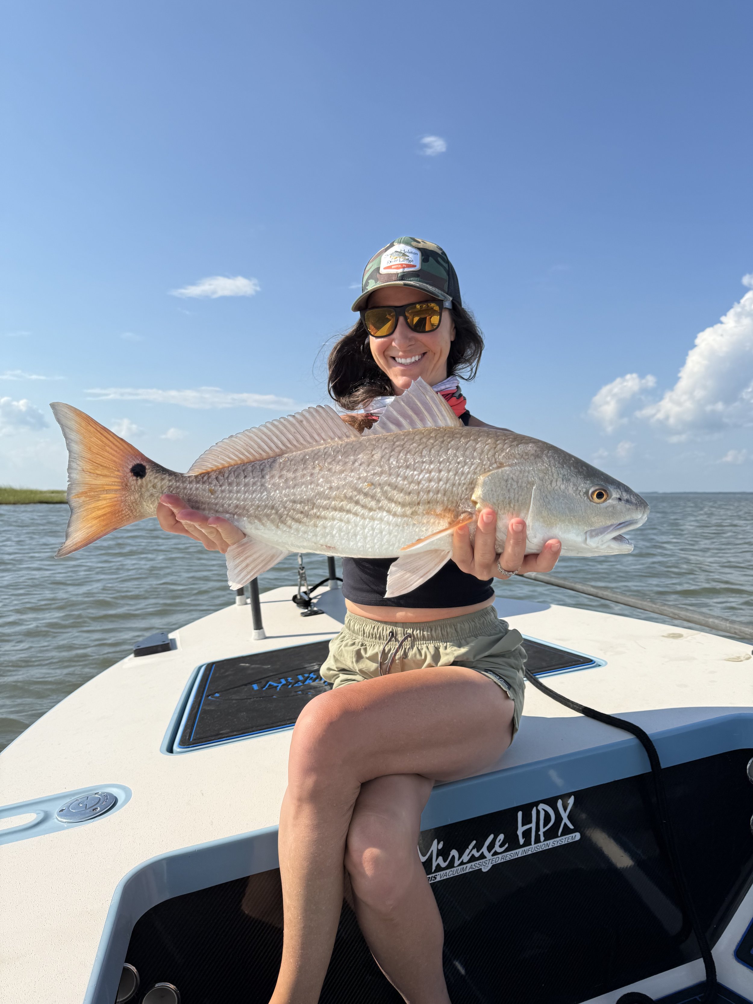 Woman sitting on a boat holding a large fish she caught, with a blue sky and water in the background.