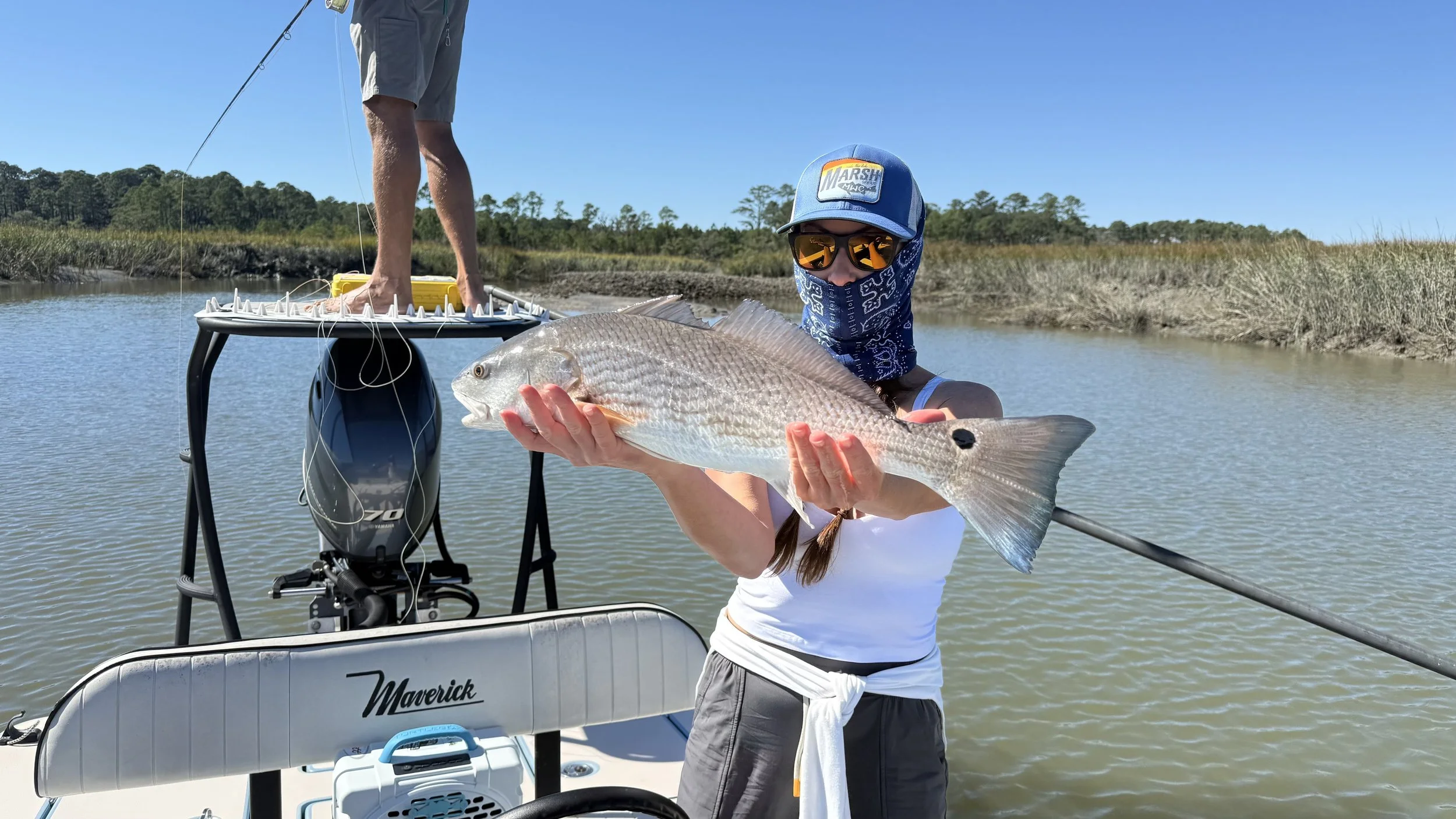 Person holding a large fish on a boat in a marsh, with another person standing on an elevated platform in the background.