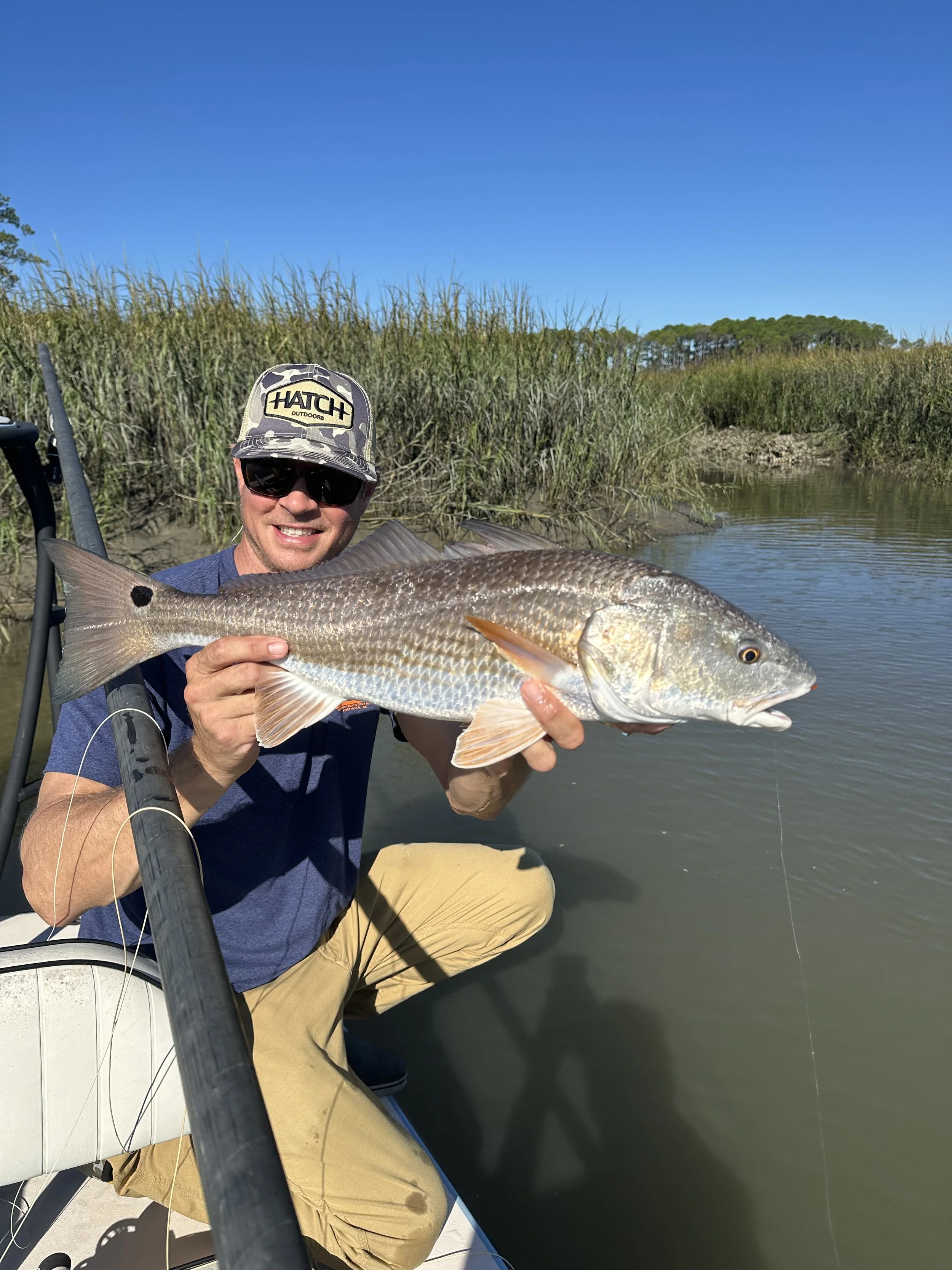 A man wearing sunglasses and a camouflage hat holding a large fish on a boat near a marshy area.