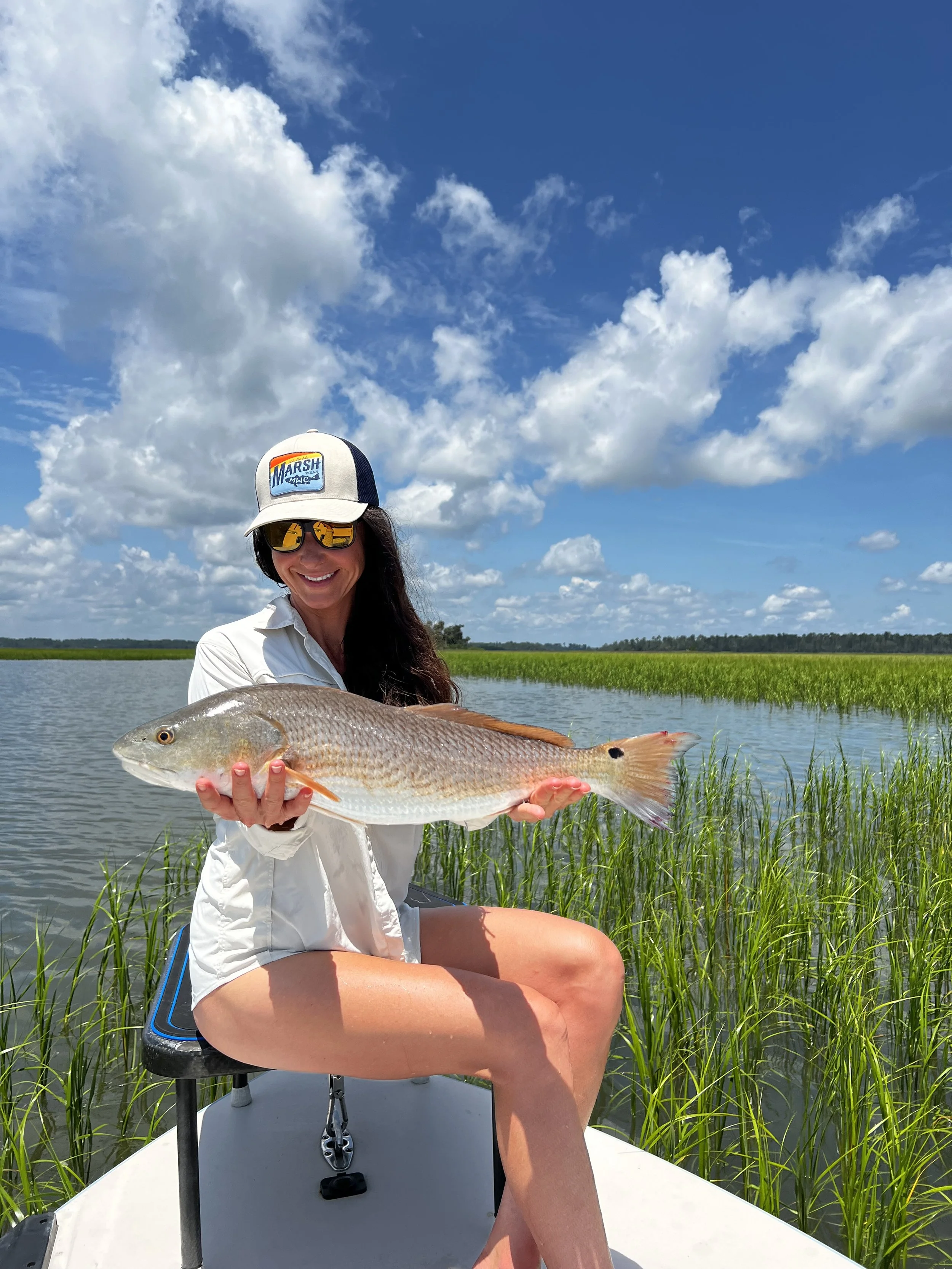 Woman sitting on a boat holding a large fish, wearing a white shirt, shorts, sunglasses, and a cap, with blue sky and clouds in the background.