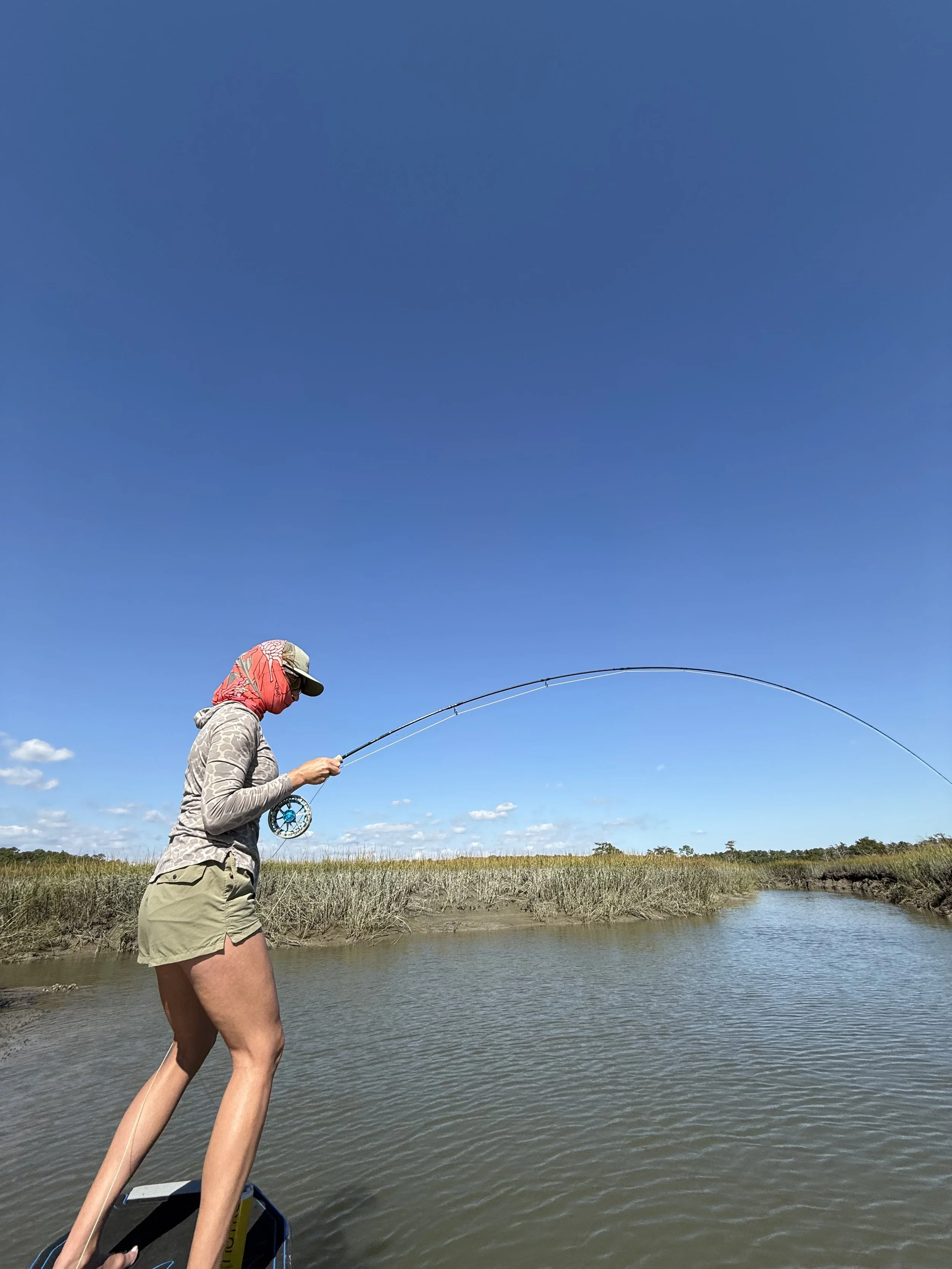Person fishing from a boat on a calm river under a clear blue sky.