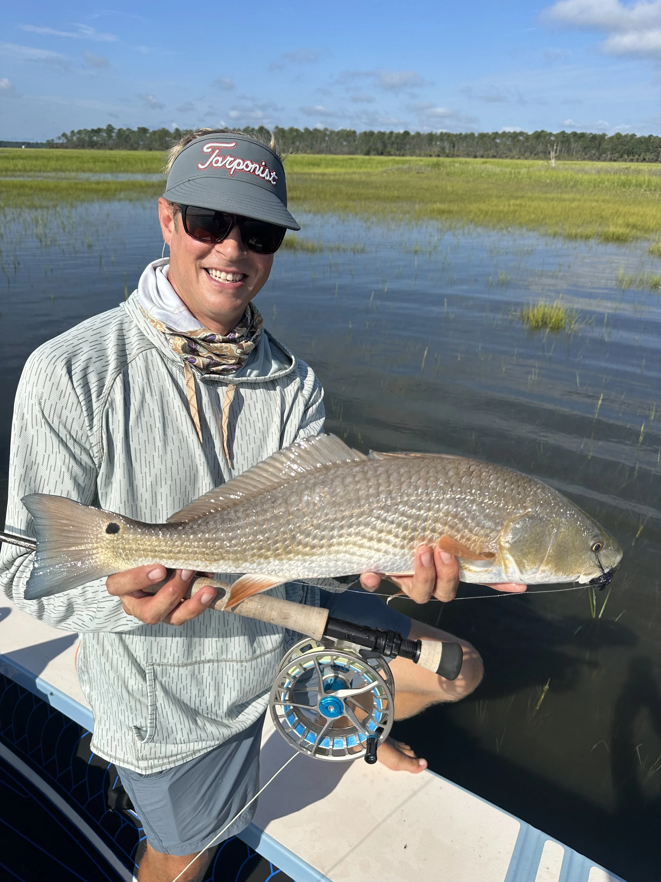 A man holding a large fish with a fishing rod in front of a water body with grassy wetlands and a blue sky.