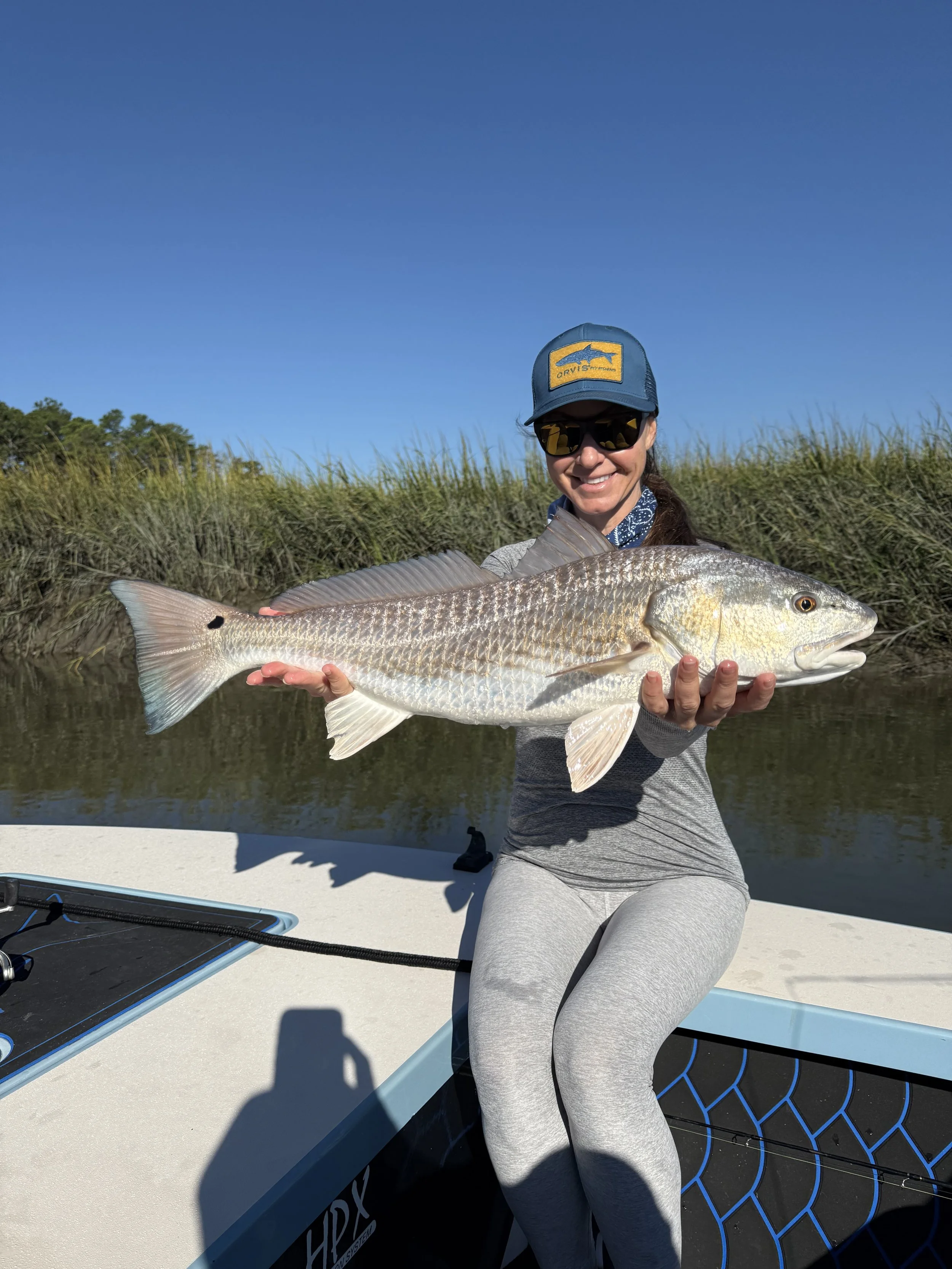 Woman sitting on a boat holding a large fish she caught, with water and grass in the background under a clear blue sky.