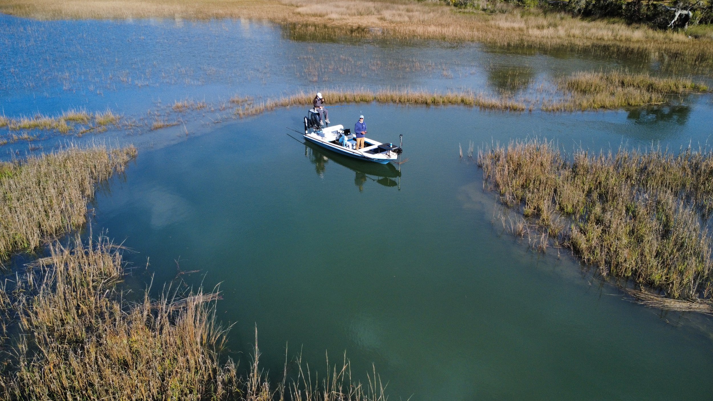 A boat with three people fishing in a marshy area with patches of tall grass and water, captured from an aerial perspective.