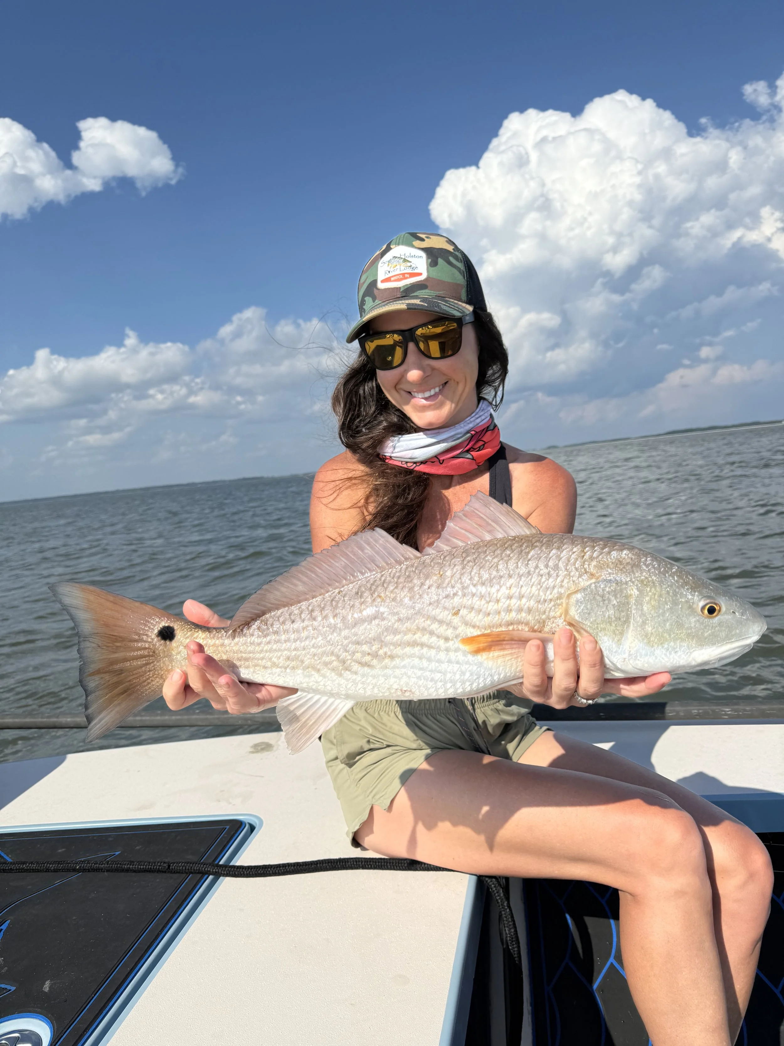 A woman wearing sunglasses, a camo hat, and a neck gaiter is sitting on a boat, smiling and holding a large fish she caught. The background shows a body of water and a partly cloudy sky.