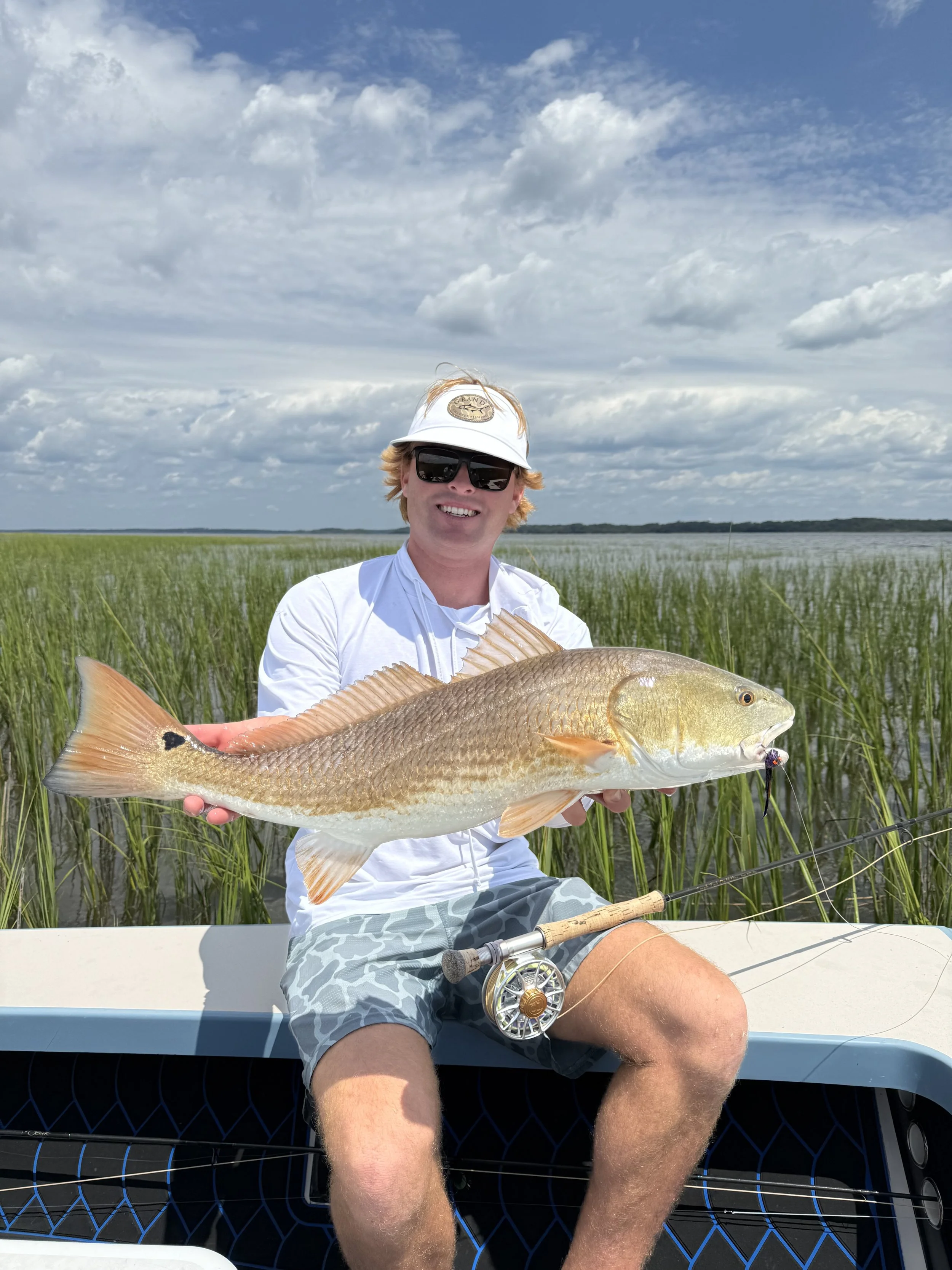 A man wearing sunglasses, a white cap, and a white shirt sitting in a boat holding a large fish with a fishing rod placed on his lap. The background features tall green marsh grass and a cloudy sky.