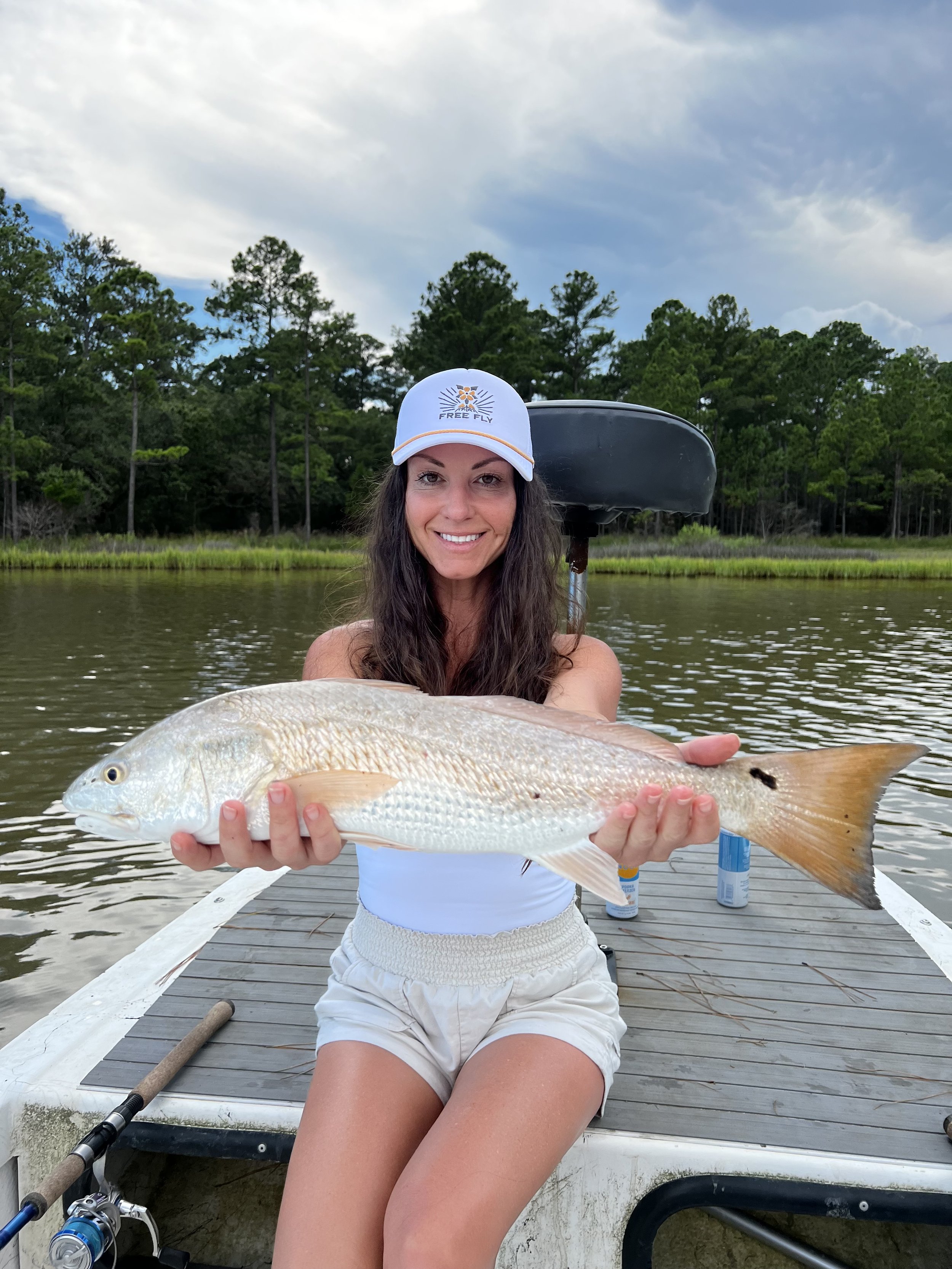 A woman in a white cap and white shorts is sitting on a wooden dock, holding a large fish with a light-colored body and a reddish tail, with a backdrop of water and green trees.