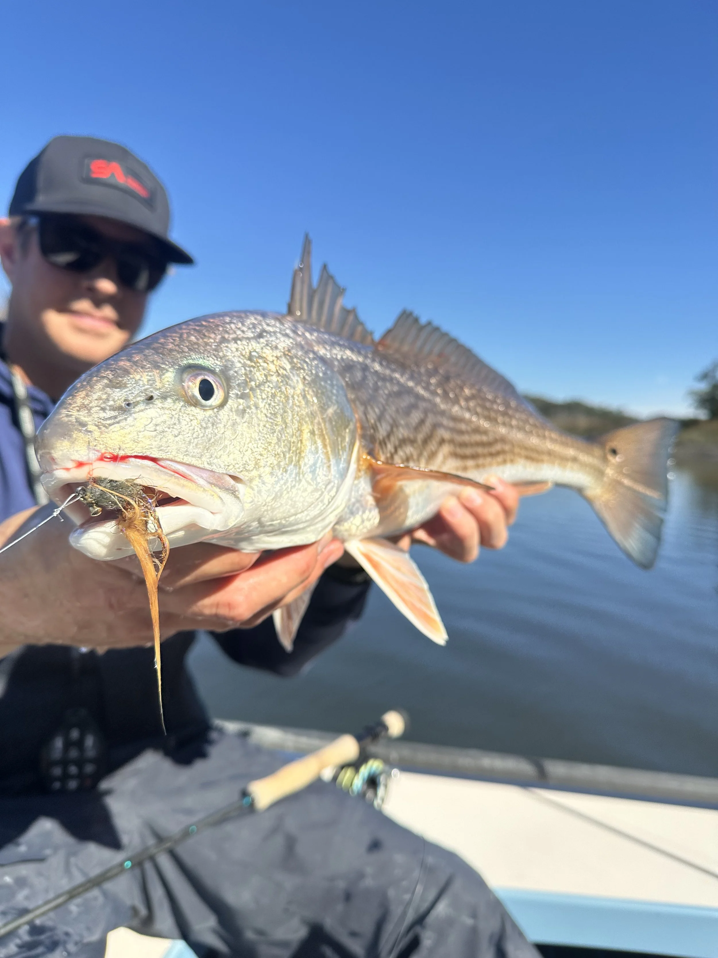 Man holding a large fish with a fishing lure in its mouth on a boat with water and blue sky in the background.