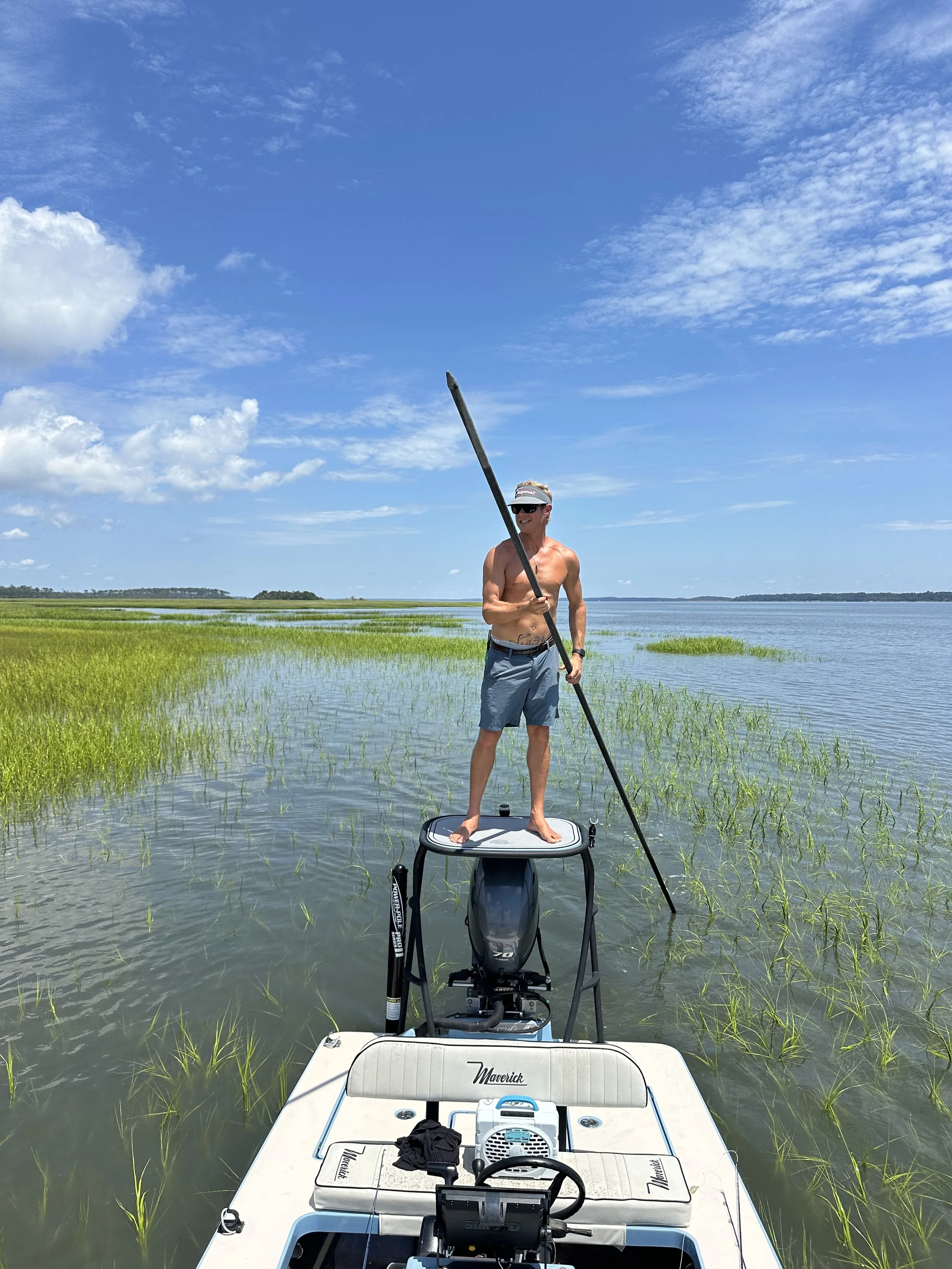 A man standing on a boat in a shallow water body, holding a pole with grass and water around him, under a partly cloudy blue sky.
