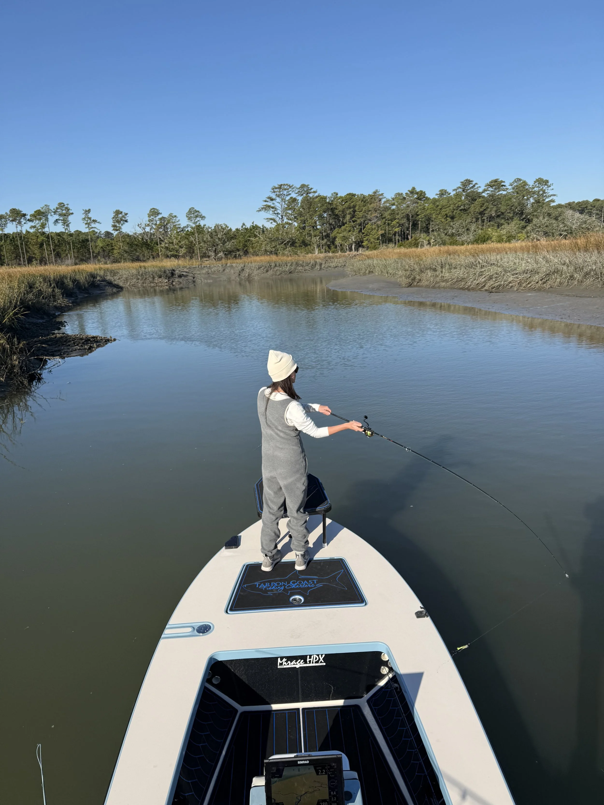 A girl fishing on a boat in a calm river with trees and marshland in the background during a clear day.