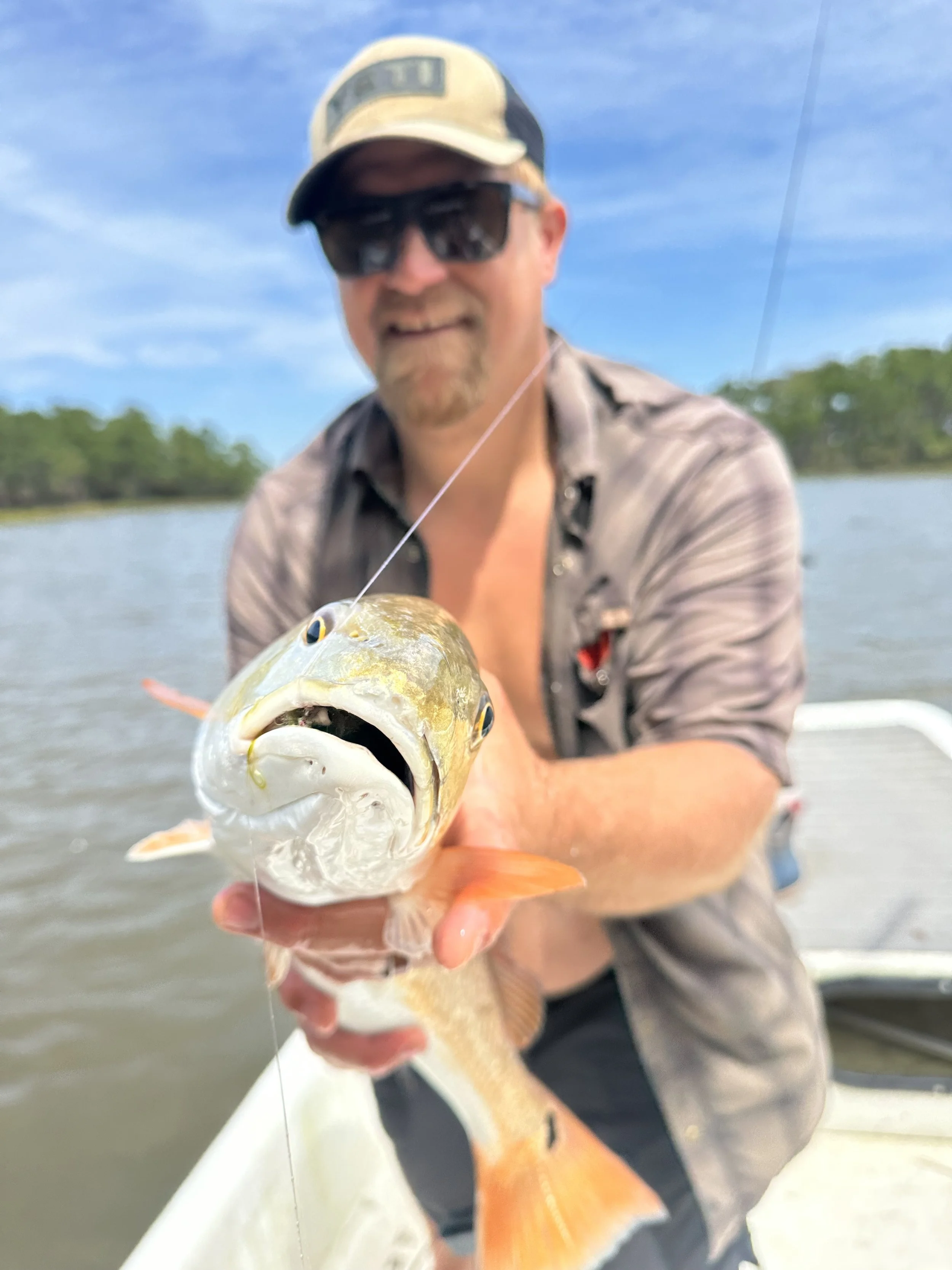 A man wearing sunglasses, a cap, and a plaid shirt holds a fish he caught on a fishing trip, standing on a boat on a river with trees and blue sky in the background.