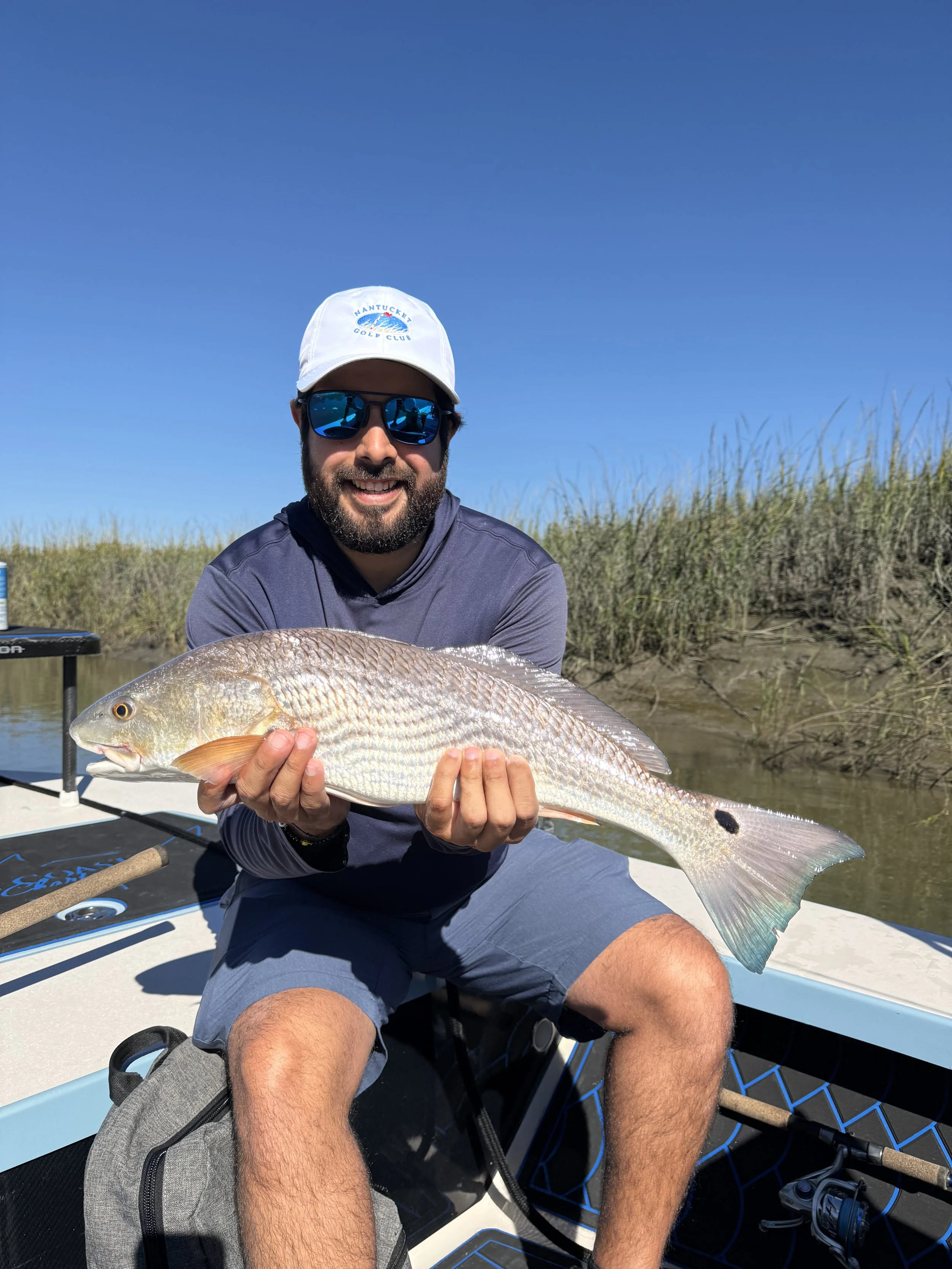 A man with a beard, wearing sunglasses, a white hat with a logo, and a gray shirt, smiling and holding a large fish on a boat near a marshy area under a clear blue sky.