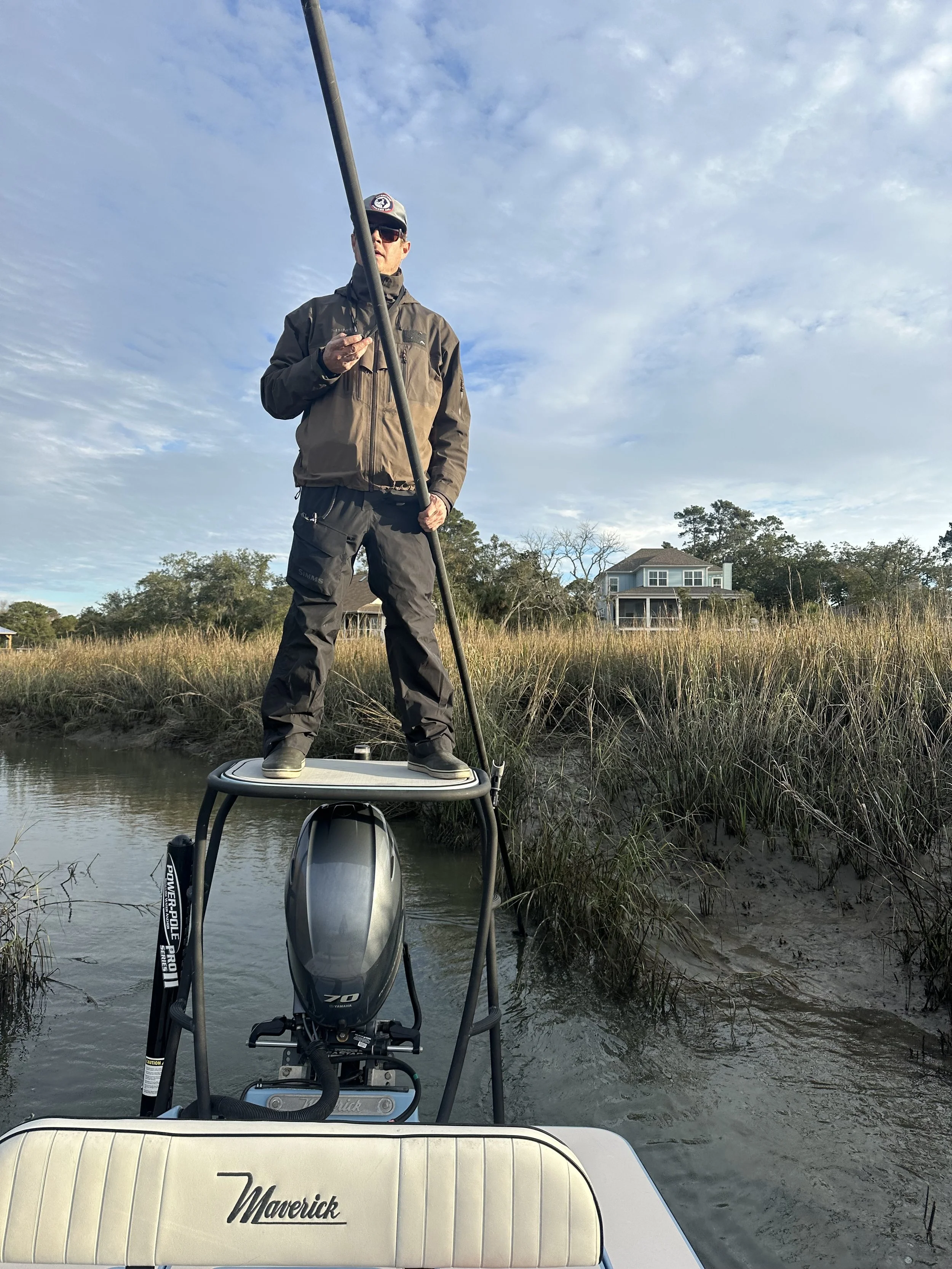 A man standing on a boat's raised platform in a marsh, holding a fishing rod and looking at his phone, with a house and trees in the background under a partly cloudy sky.