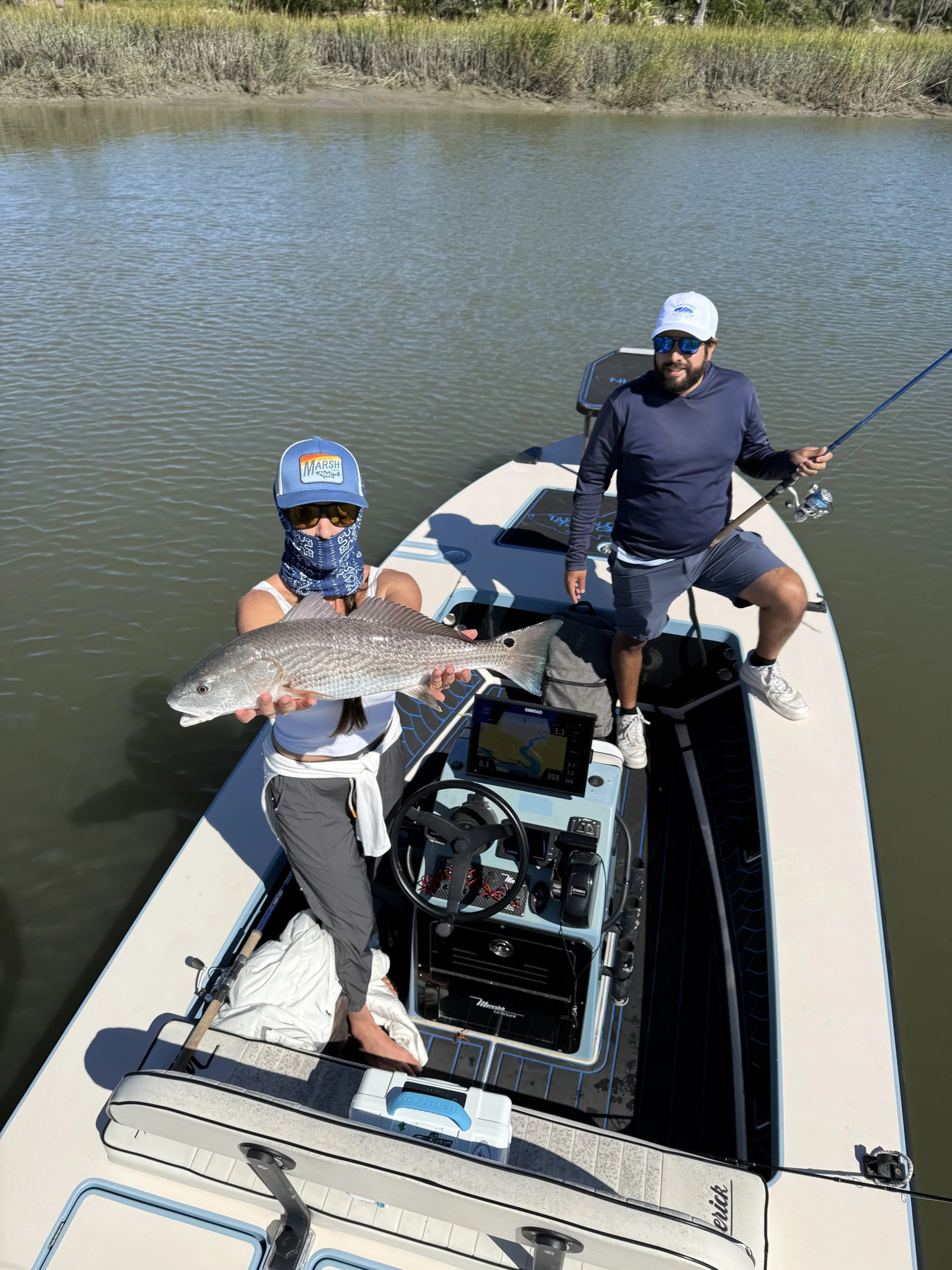 Two men on a boat in a lake, one holding a large fish, with fishing gear and navigation equipment visible.