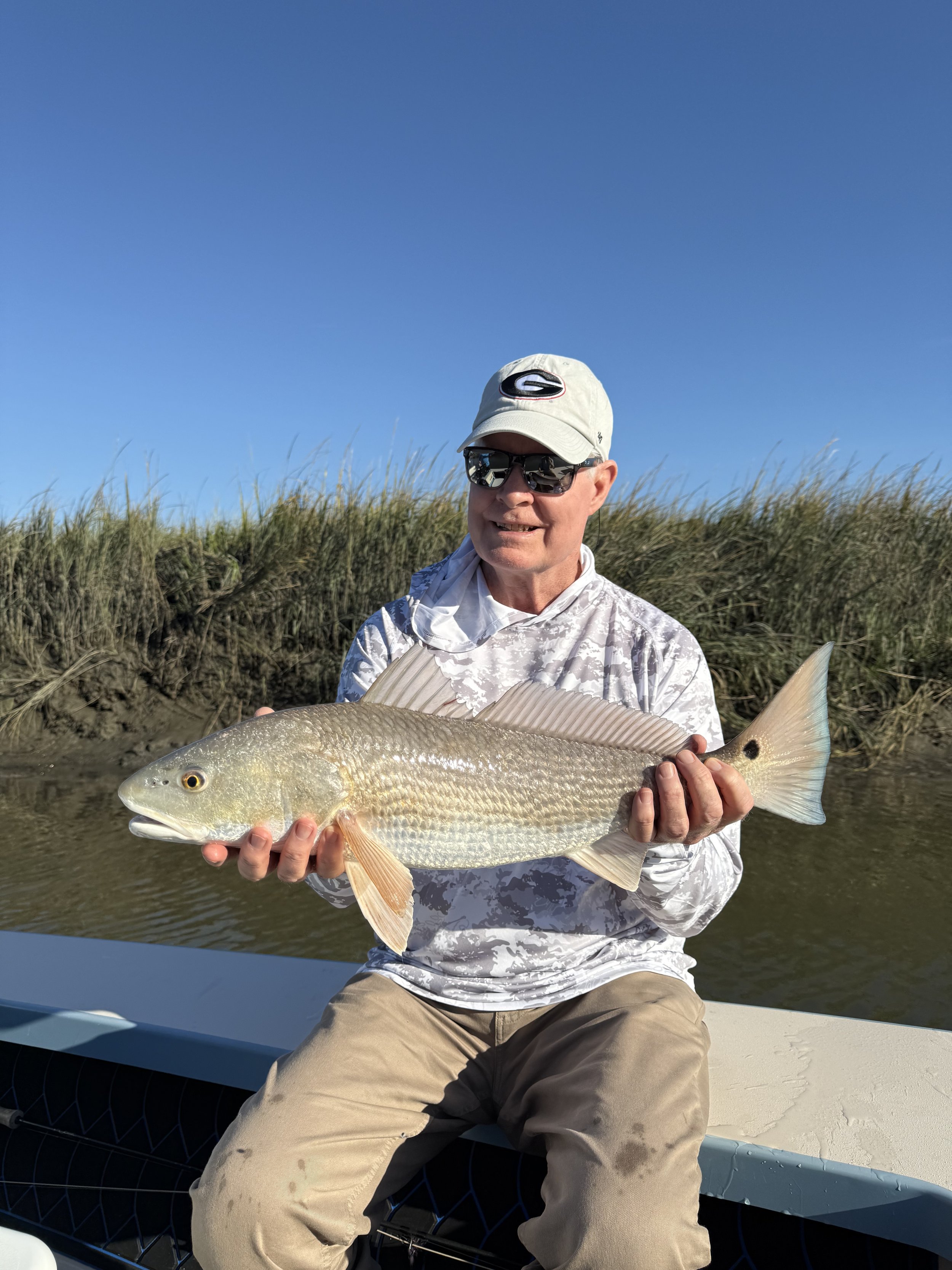 Man wearing sunglasses and a cap holding a large fish on a boat near grassy shoreline under clear blue sky.