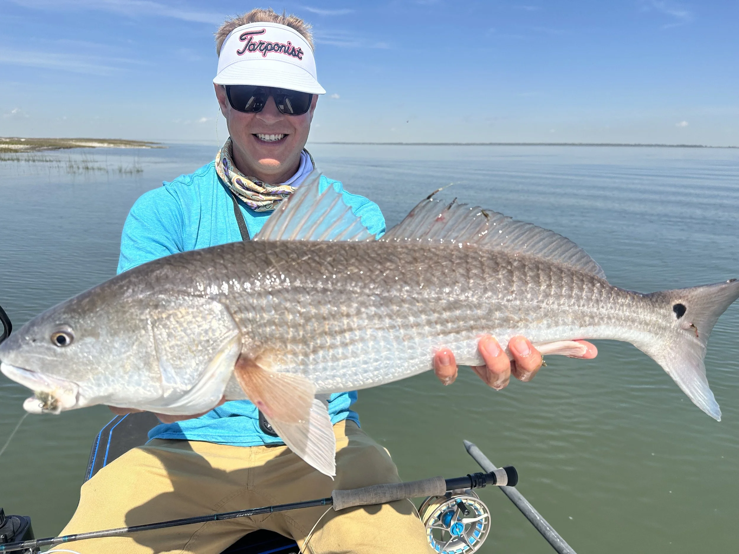 A man wearing sunglasses, a white visor with 'Tarponic' written on it, a blue long-sleeve shirt, and tan pants, is smiling while holding a large red drum fish over a body of water.