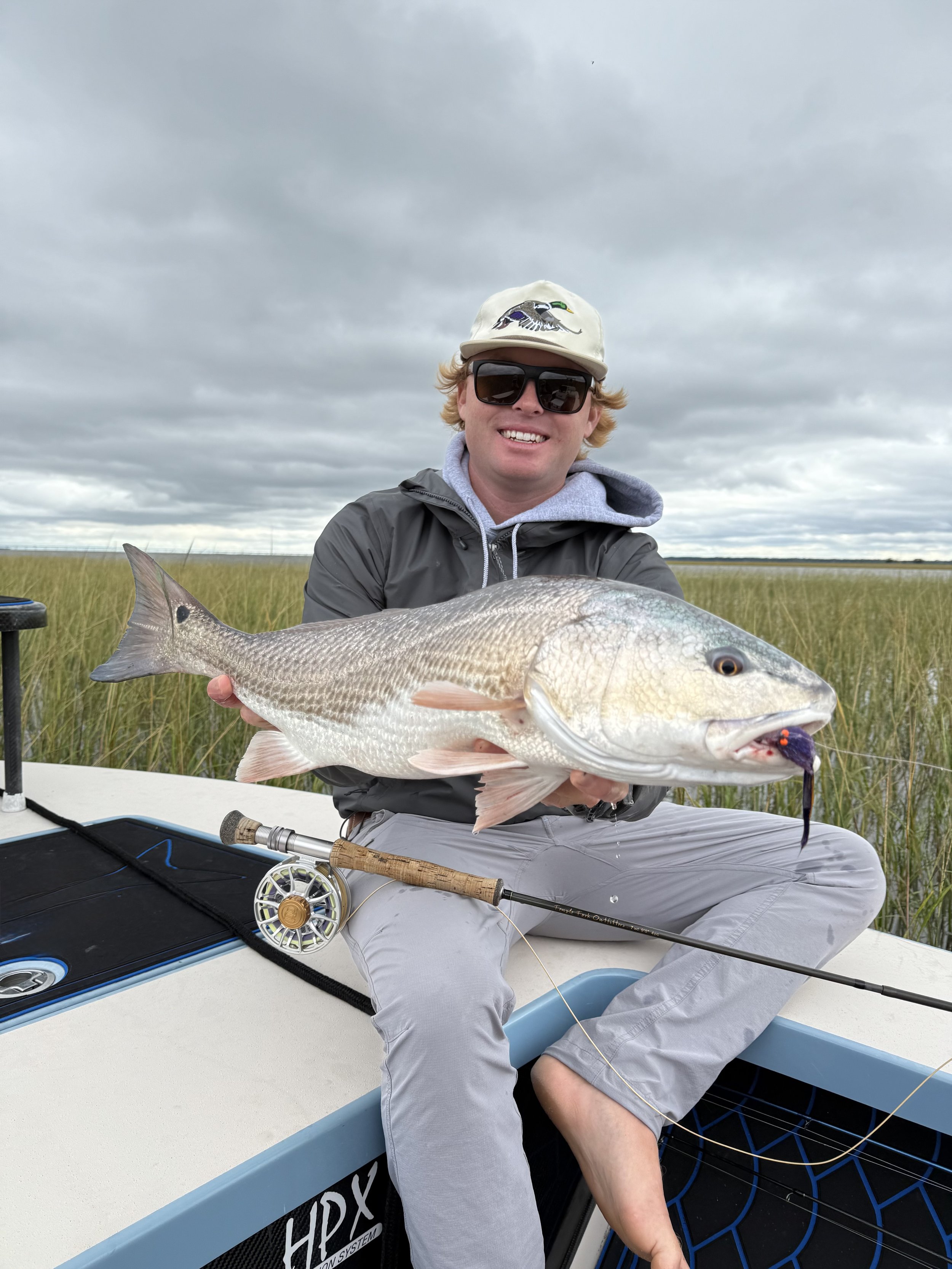 A young man with sunglasses, a cap, and a gray hoodie sitting on a boat, holding a large fish with a fishing rod nearby, in a marshy area with cloudy skies.