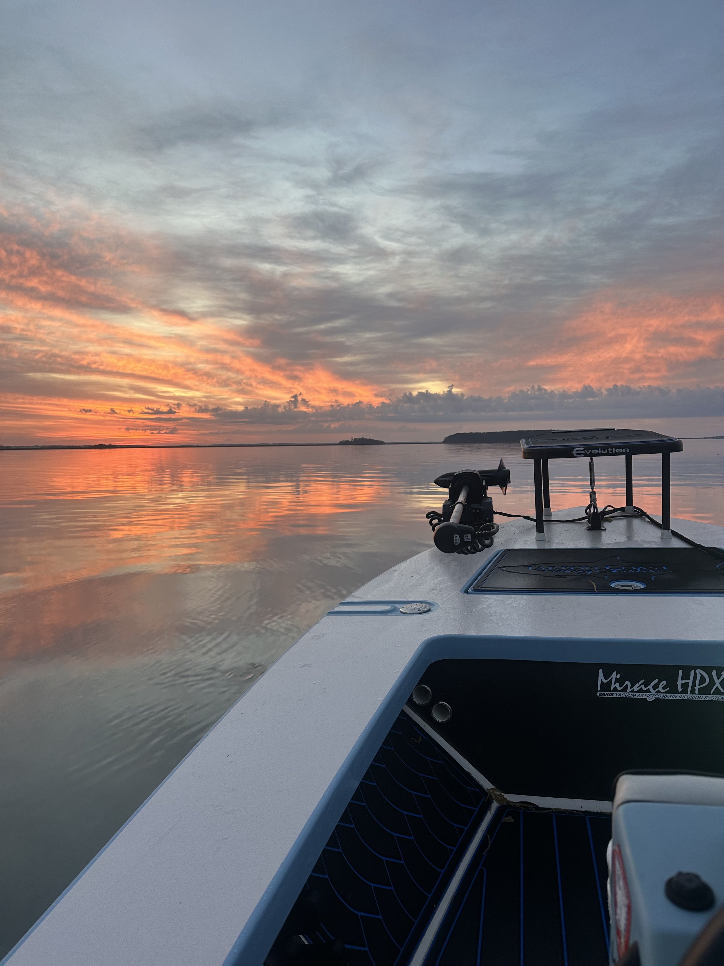 View from a boat during sunset with calm water reflecting the colorful sky, and part of the boat's stern, including fishing gear and a solar panel.