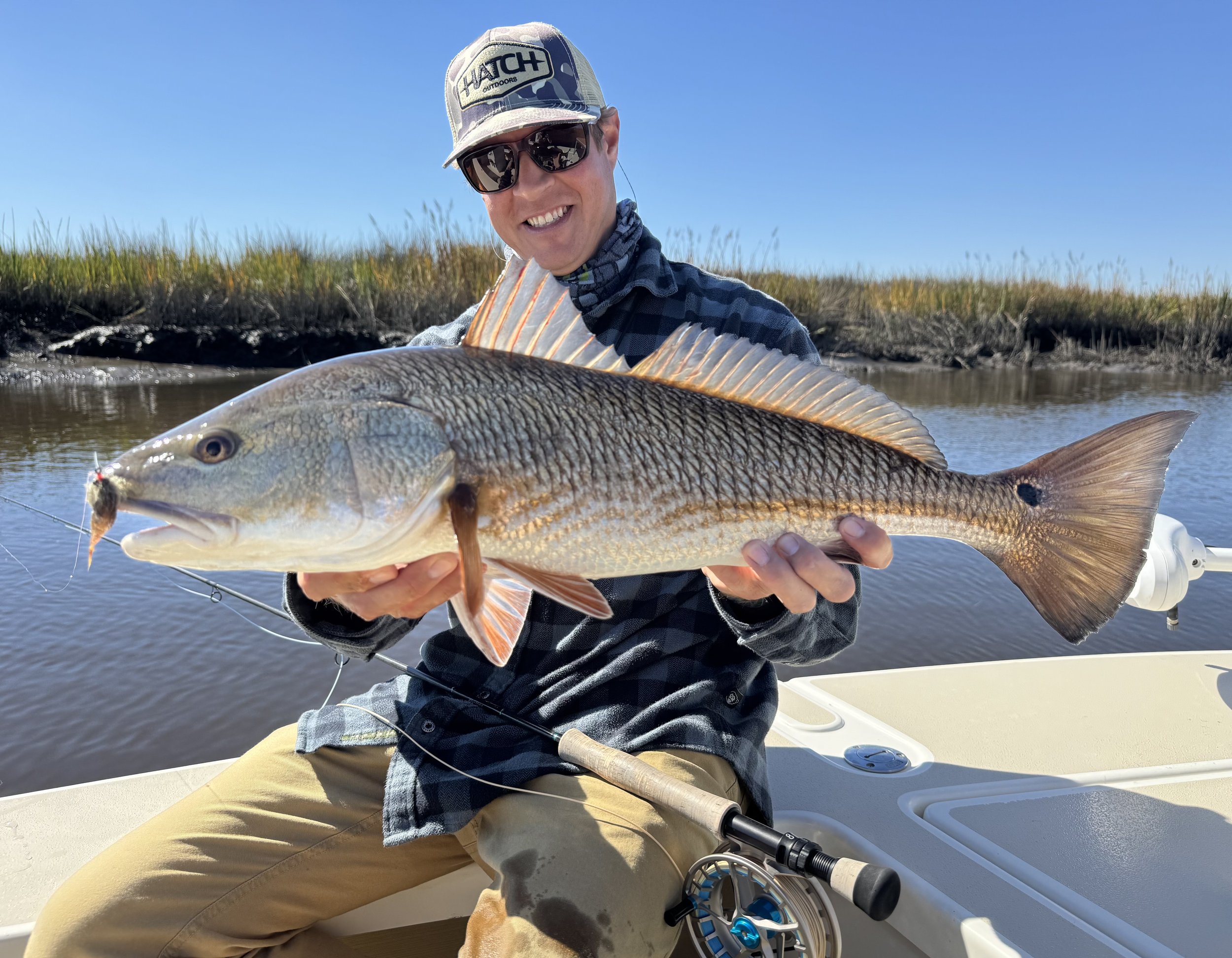 A man in a plaid shirt, wearing sunglasses and a cap, smiling and holding a large redfish on a boat, with water and marshy land in the background on a clear day.