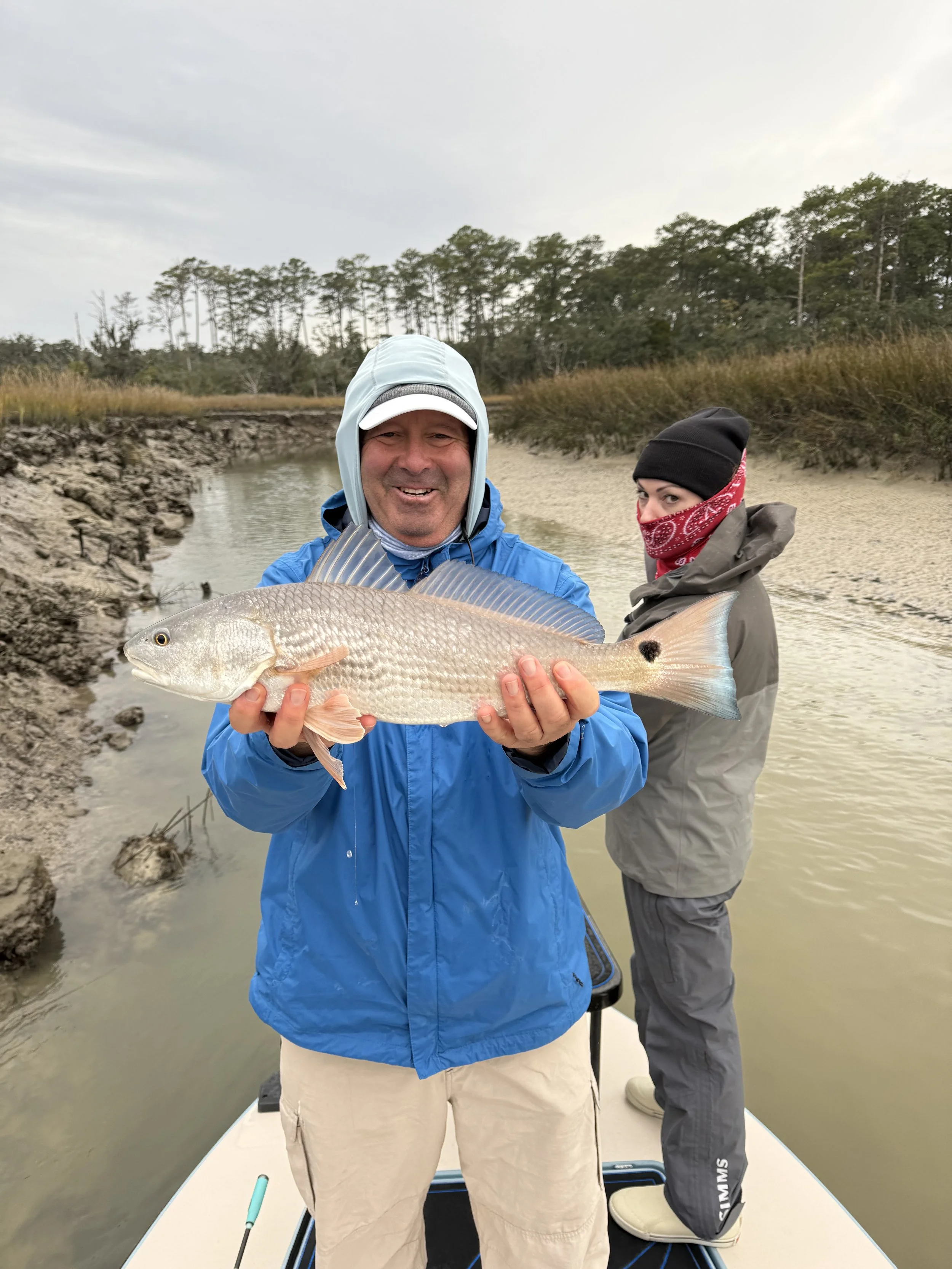 Two people on a boat in a marsh holding a large fish, with trees and grassy marshland in the background.