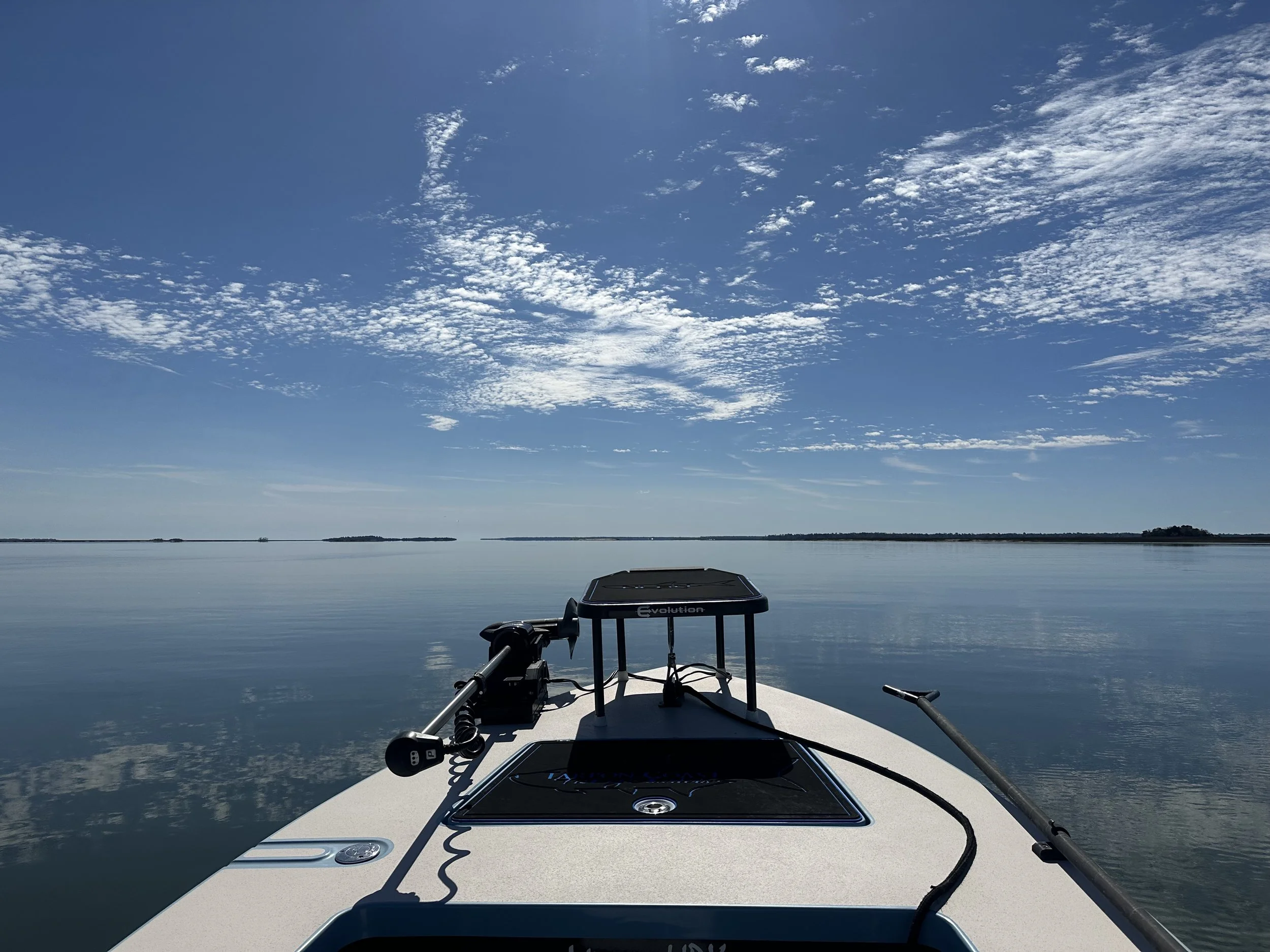 View from a boat on a calm lake with a clear blue sky and scattered clouds, showing the boat's front and fishing equipment.