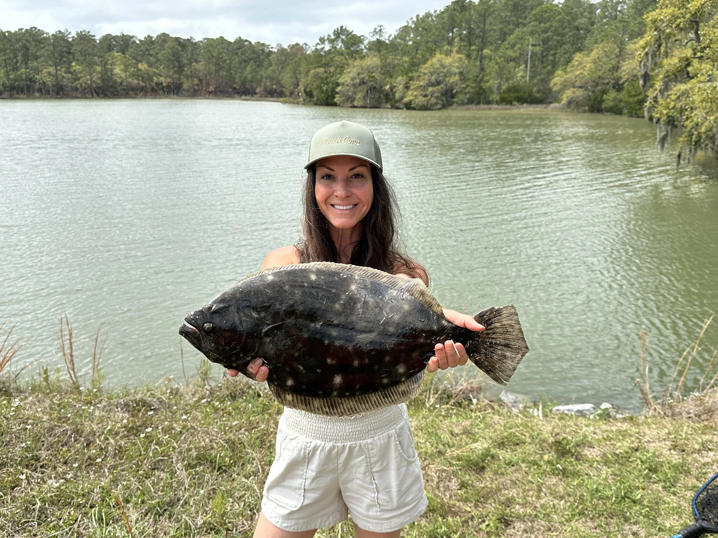 A woman with long dark hair wearing a green hat and white shorts, standing near a lake, holding a large black fish and smiling.