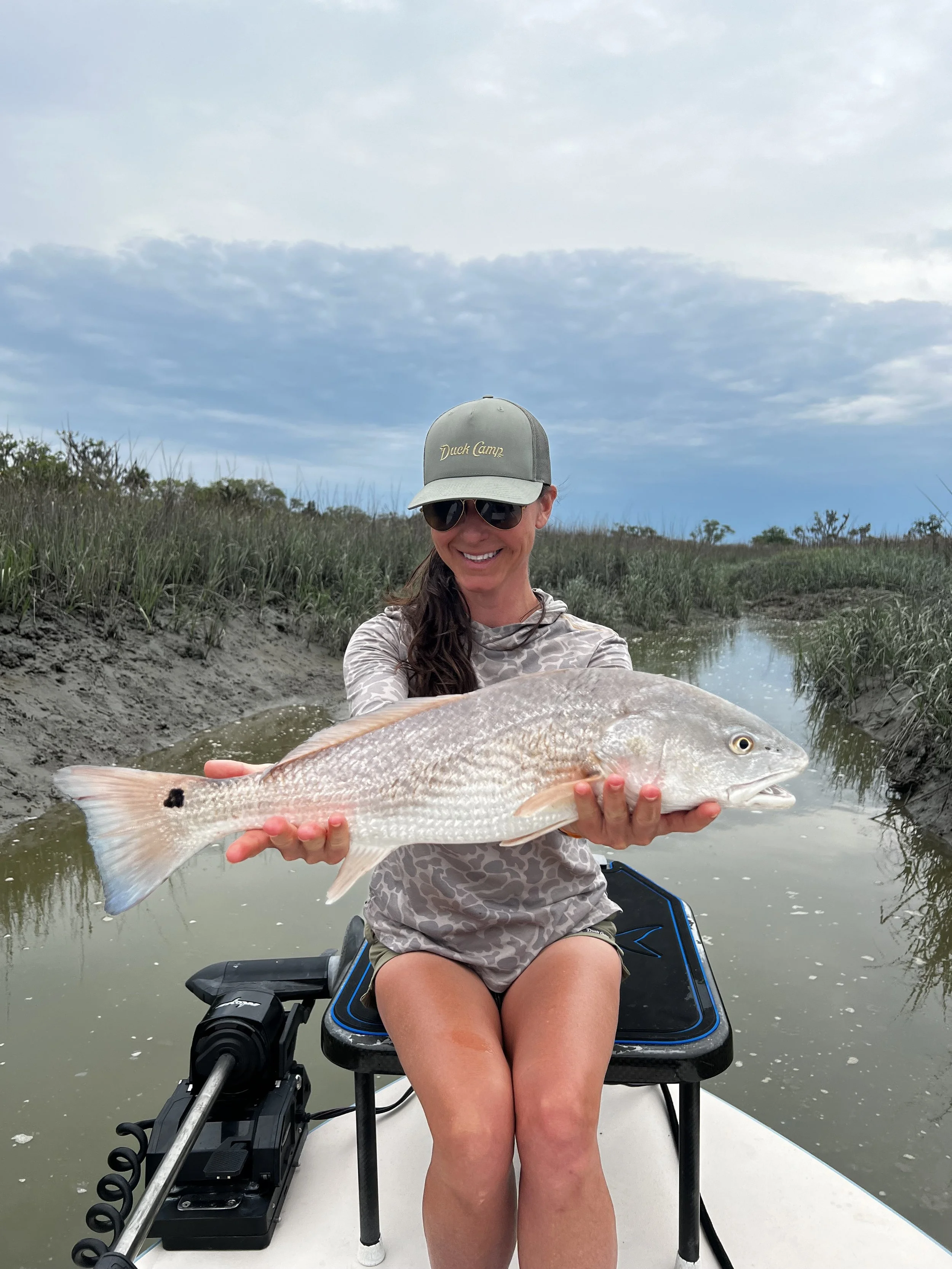 A woman sitting on a boat holding a large fish with a marshy wetlands background and cloudy sky.
