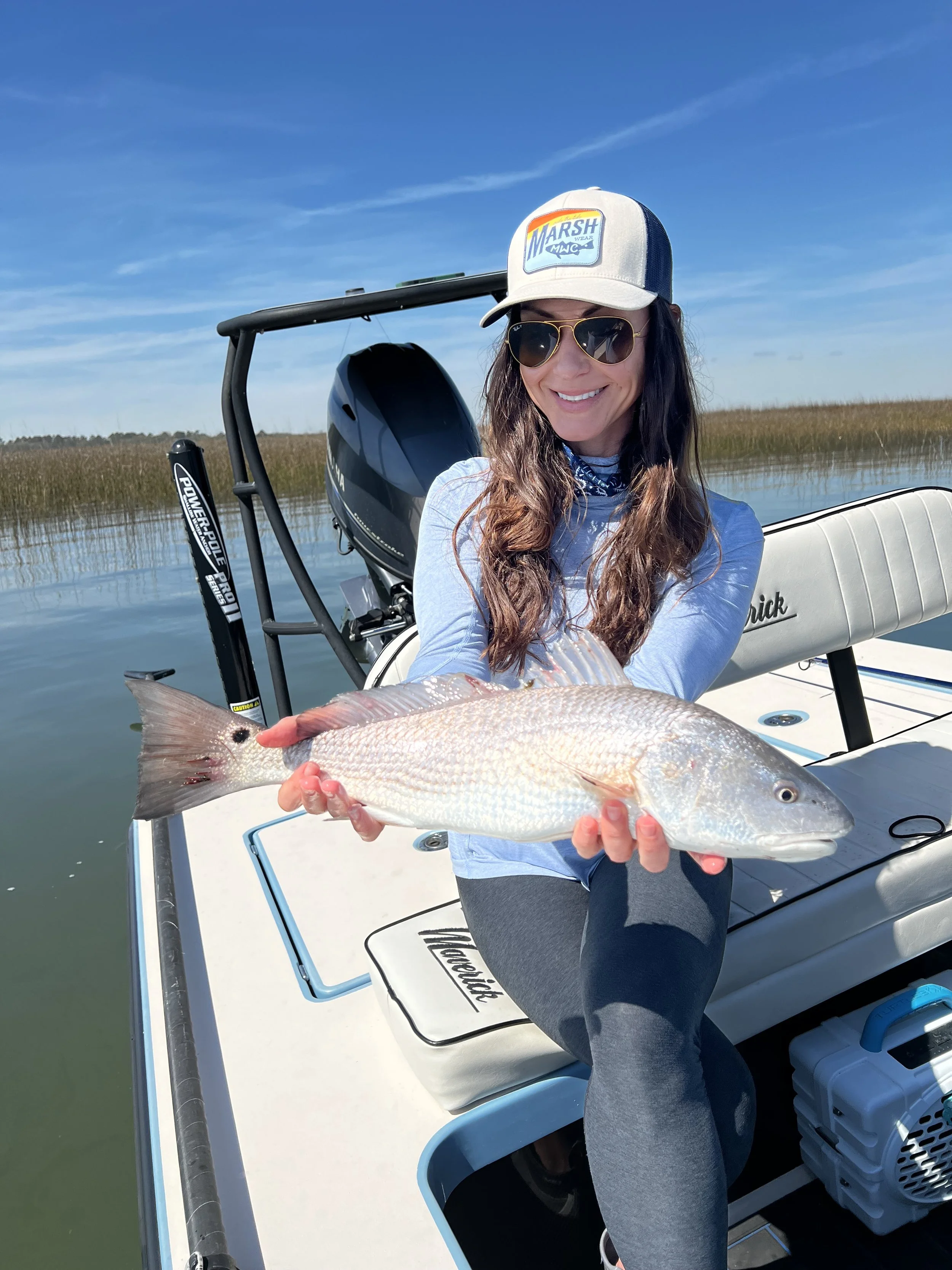 Woman in a boat holding a large fish, wearing sunglasses and a trucker hat, with a body of water and marsh grass in the background on a sunny day.