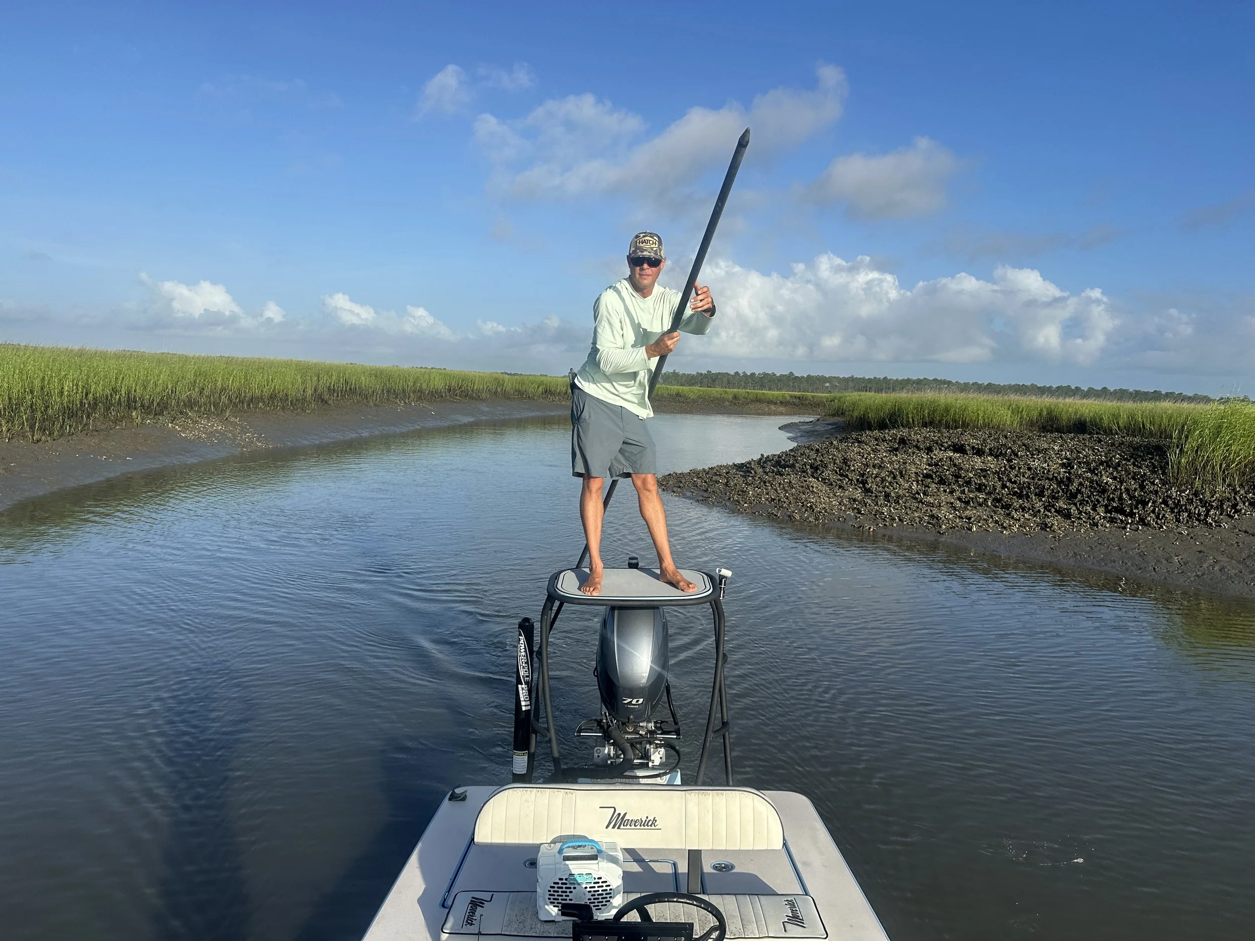 A man standing on the bow of a boat, holding a paddle, in a river surrounded by marshland with green grass and a partly cloudy sky.