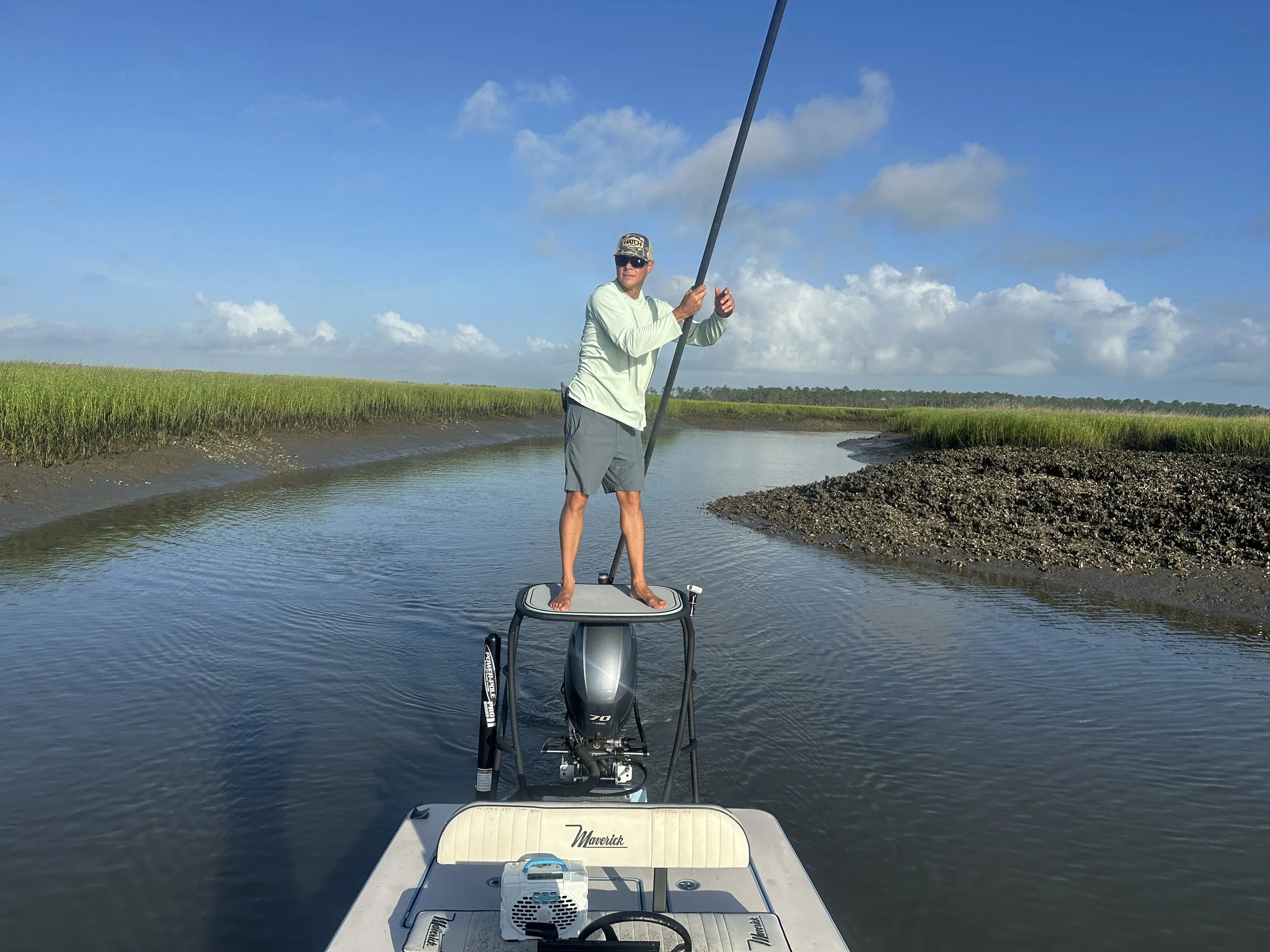 Man standing on a boat holding a fishing pole on a calm waterway surrounded by grassy banks under a partly cloudy sky.