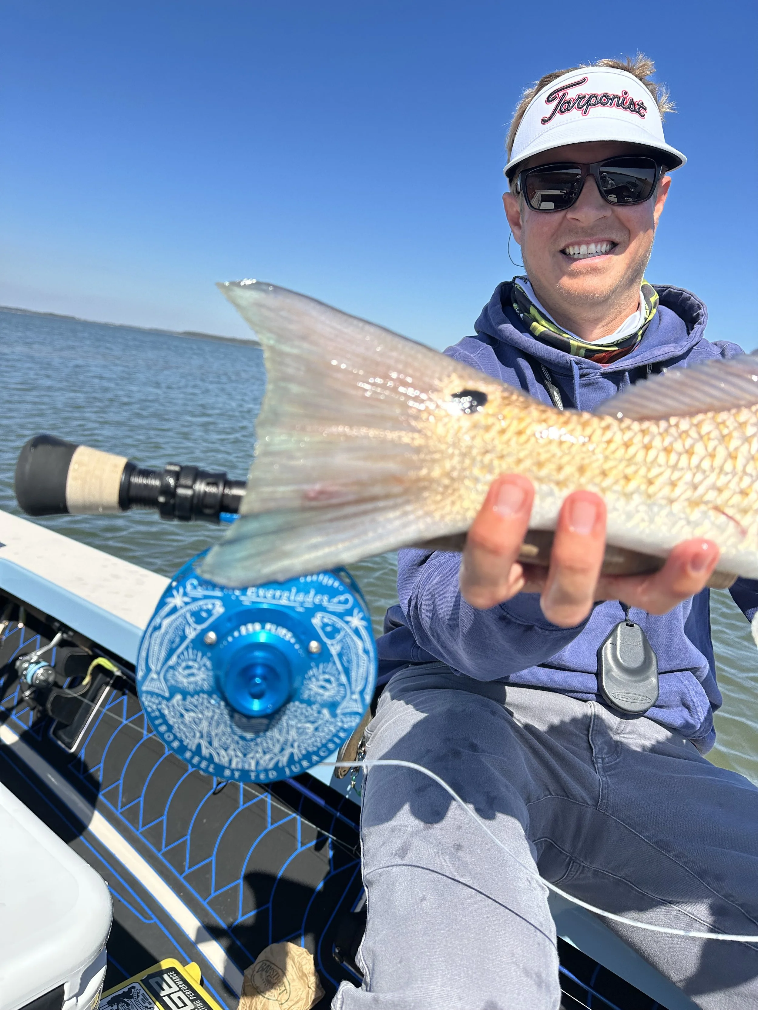 A man wearing sunglasses and a white visor hat smiling while holding a large fish on a boat.
