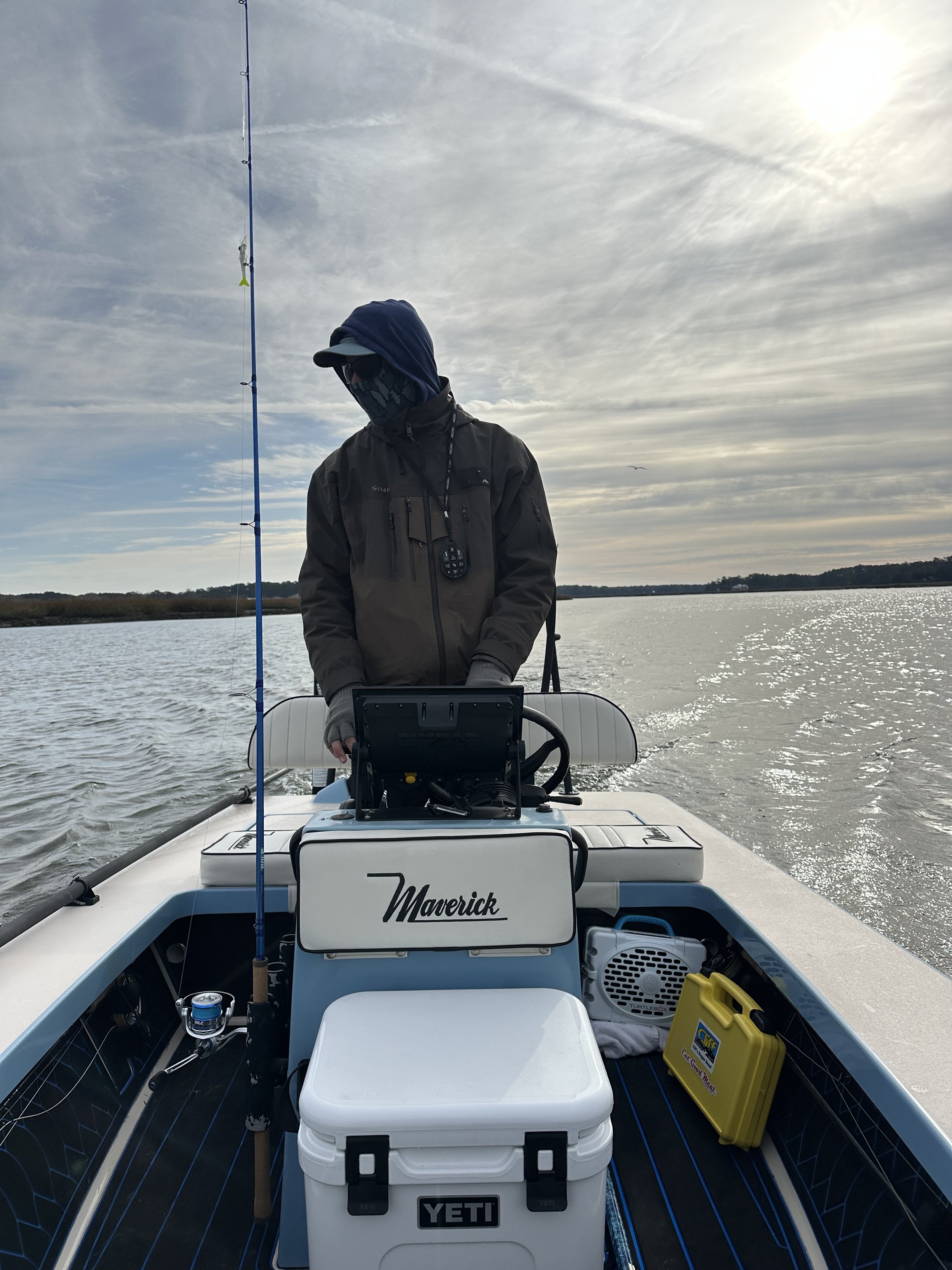 Person fishing on a boat on a lake in overcast weather, wearing a jacket, hoodie, and sunglasses.